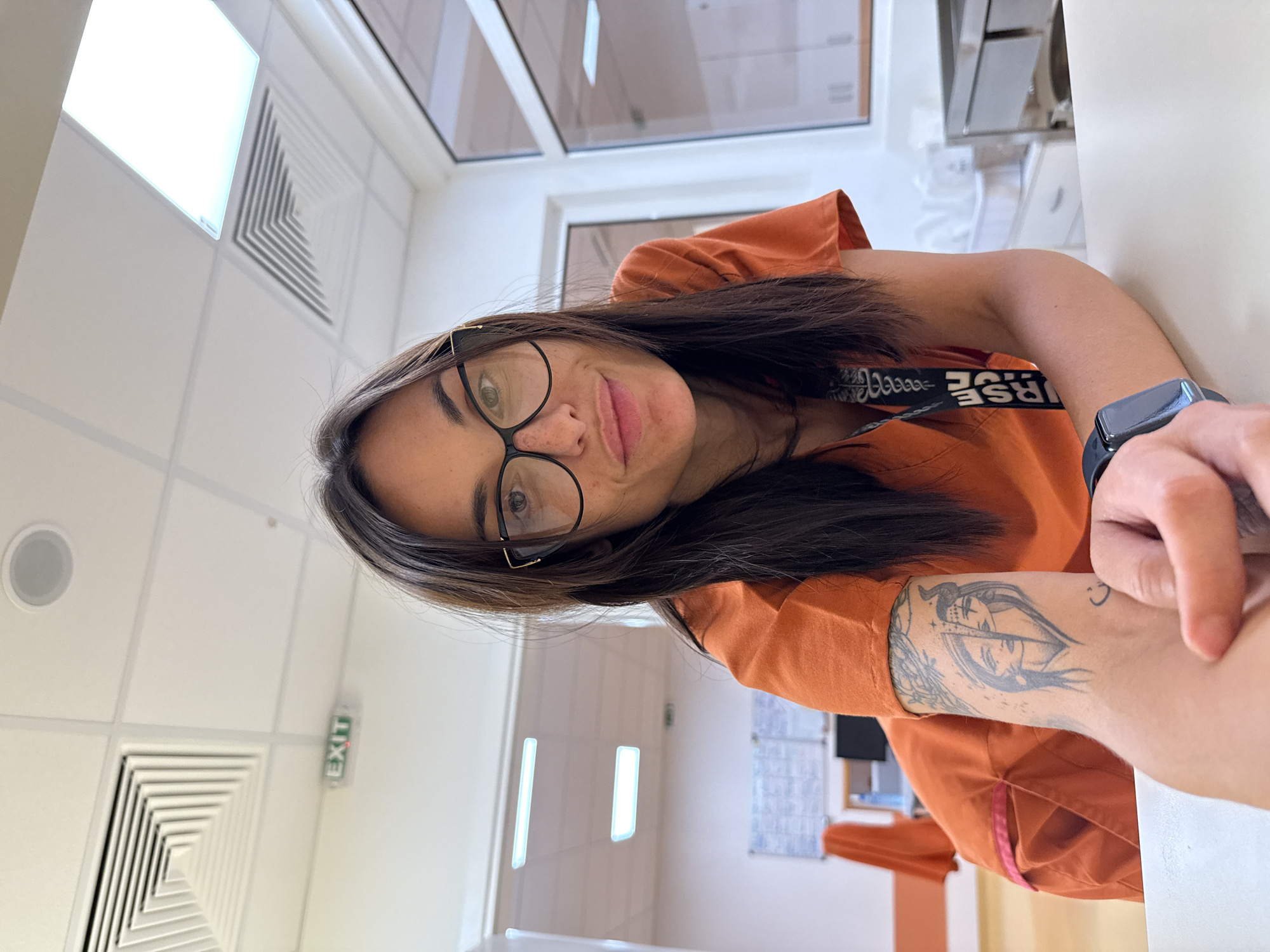 A young woman with dark hair and glasses, wearing an orange scrub top, poses for a selfie in what appears to be a medical facility. She has a tattoo on her left arm. An "EXIT" sign is visible on the ceiling in the background. The environment includes white ceiling tiles, fluorescent lighting, and what looks like a reception desk or counter. The scene does not contain any information that would indicate the location in the city of Pleven, Bulgaria.