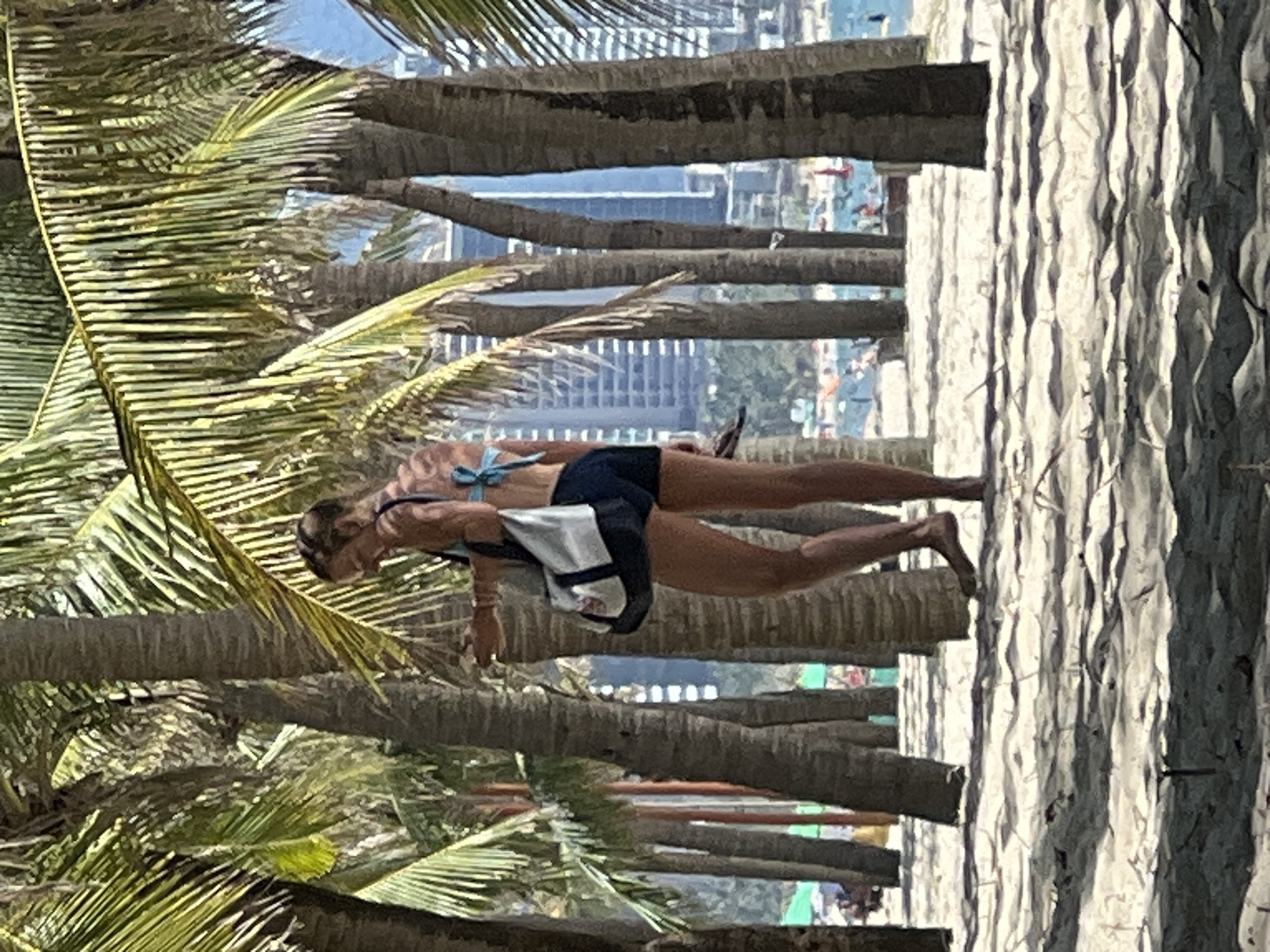 A woman walks along a sandy beach lined with palm trees, with a city skyline in the background. The woman is wearing a blue bikini top and dark shorts, and is carrying a bag. She has a tan that appears to be peeling on her back. In the distance, several buildings are visible, suggesting an urban setting. The beach also shows signs of other people, with some figures visible near the water. The overall scene is consistent with a tropical or subtropical coastal area. The presence of palm trees and a prominent city skyline suggests the location could be a popular tourist spot within a major coastal city. Based on the provided context, the location is Đà Nẵng, Vietnam, a well-known coastal city.