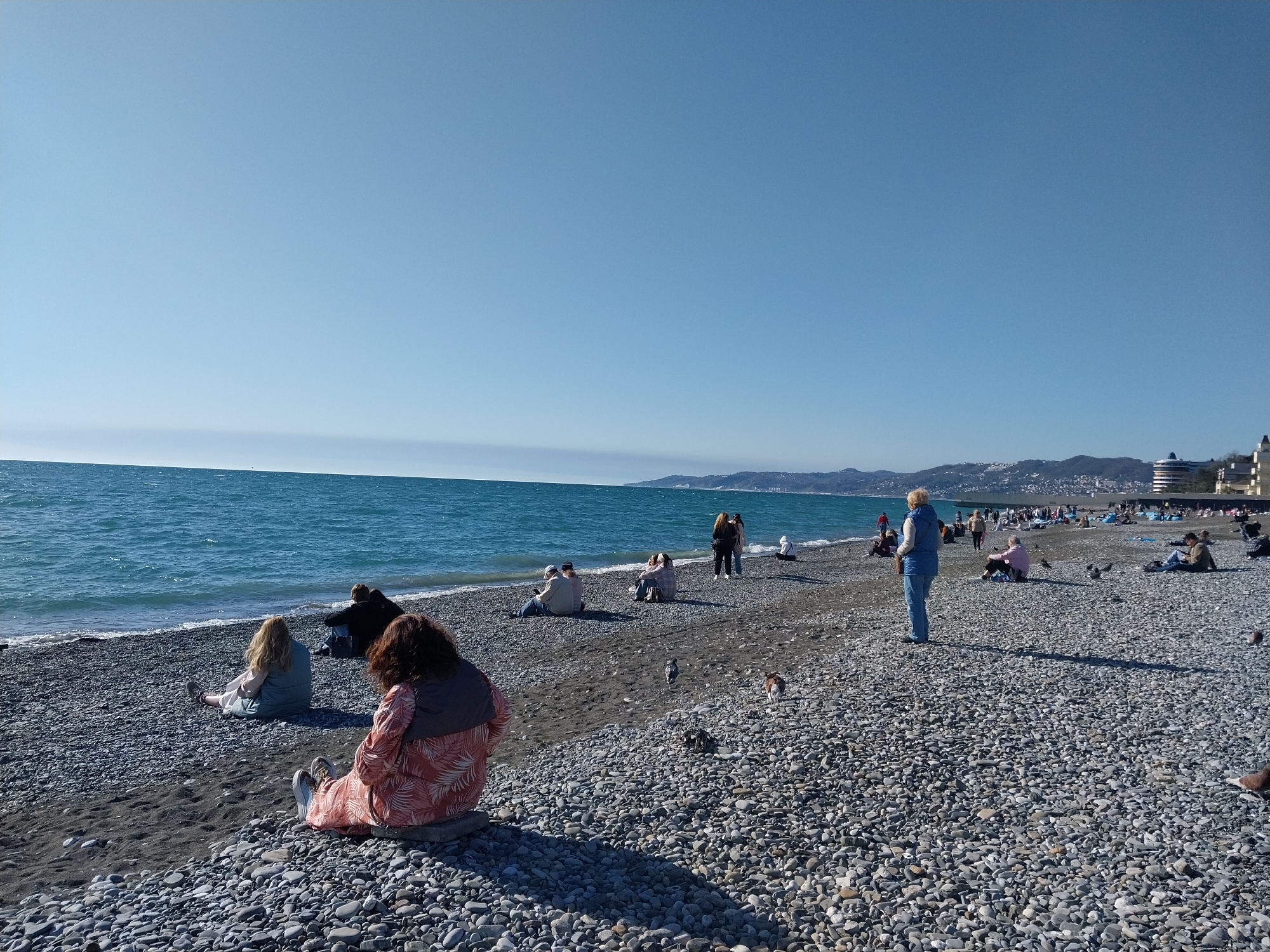 The image depicts a sunny day at a pebble beach in Gornoye Loo, Russia, bordering the Black Sea. The scene features a wide expanse of calm, blue-green water meeting a shore covered in grey and dark pebbles. A clear, light blue sky dominates the upper portion of the frame, with distant mountains and some coastal buildings visible along the horizon to the right.
Numerous individuals are scattered across the beach, engaged in leisurely activities. Many people are seated directly on the pebbles, some facing the sea, others in small groups, possibly conversing. In the foreground, a person with dark hair wearing a distinctive pink and white patterned top is seated with their back to the camera, next to another person with blonde hair in a light blue top. Further back, a person in a blue jacket and jeans stands, looking towards the water. Several other figures are seen sitting, standing, or walking along the shoreline. A few pigeons are visible on the beach among the pebbles.
The time of day appears to be late morning or early afternoon, indicated by the bright sunlight and visible shadows cast by the individuals. The weather is clear and pleasant, suggesting a calm and relaxed atmosphere. No discernible text is visible within the image.