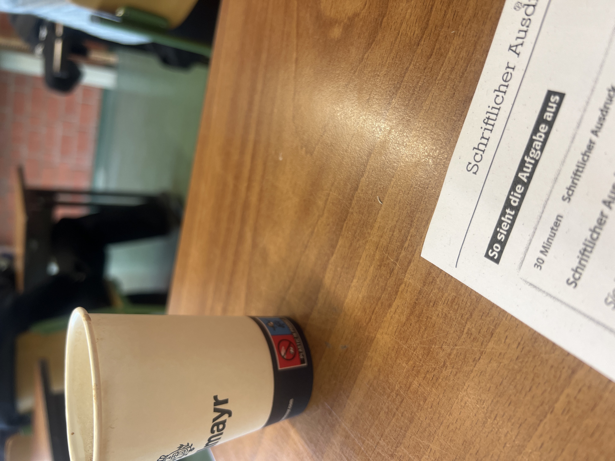 The image shows a desk with a coffee cup from "mayr" brand and a document with the title "Schriftlicher Ausd". The document also has "So sieht die Aufgabe aus" and "30 Minuten Schriftlicher Ausdruck" written on it. The desk is made of wood. The background shows a blurry view of what appears to be a room with chairs and possibly a window. The picture was taken in an unspecified indoor location; the image provides no information about the city of Bayreuth, Germany.