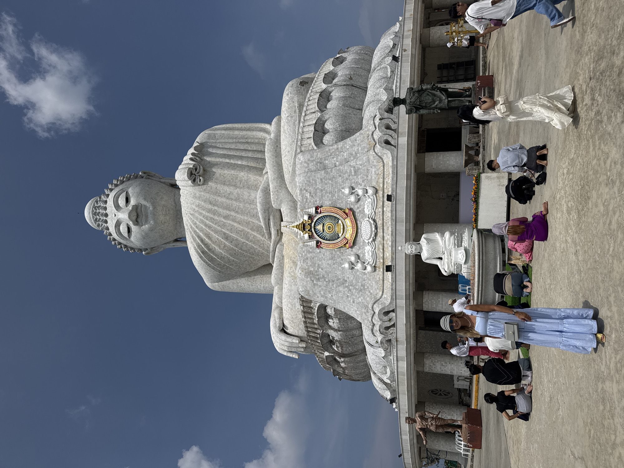 The image depicts the Big Buddha statue in Karon, Thailand. It's a large, white marble Buddha statue, with a clear blue sky in the background and some clouds. The statue is elevated on a platform, and several people are observing it. Some of them are kneeling or praying in front of it. A few statues are visible around the Buddha statue. Some visitors are seen taking photos. The image is taken from a low angle, making the Buddha statue appear massive.