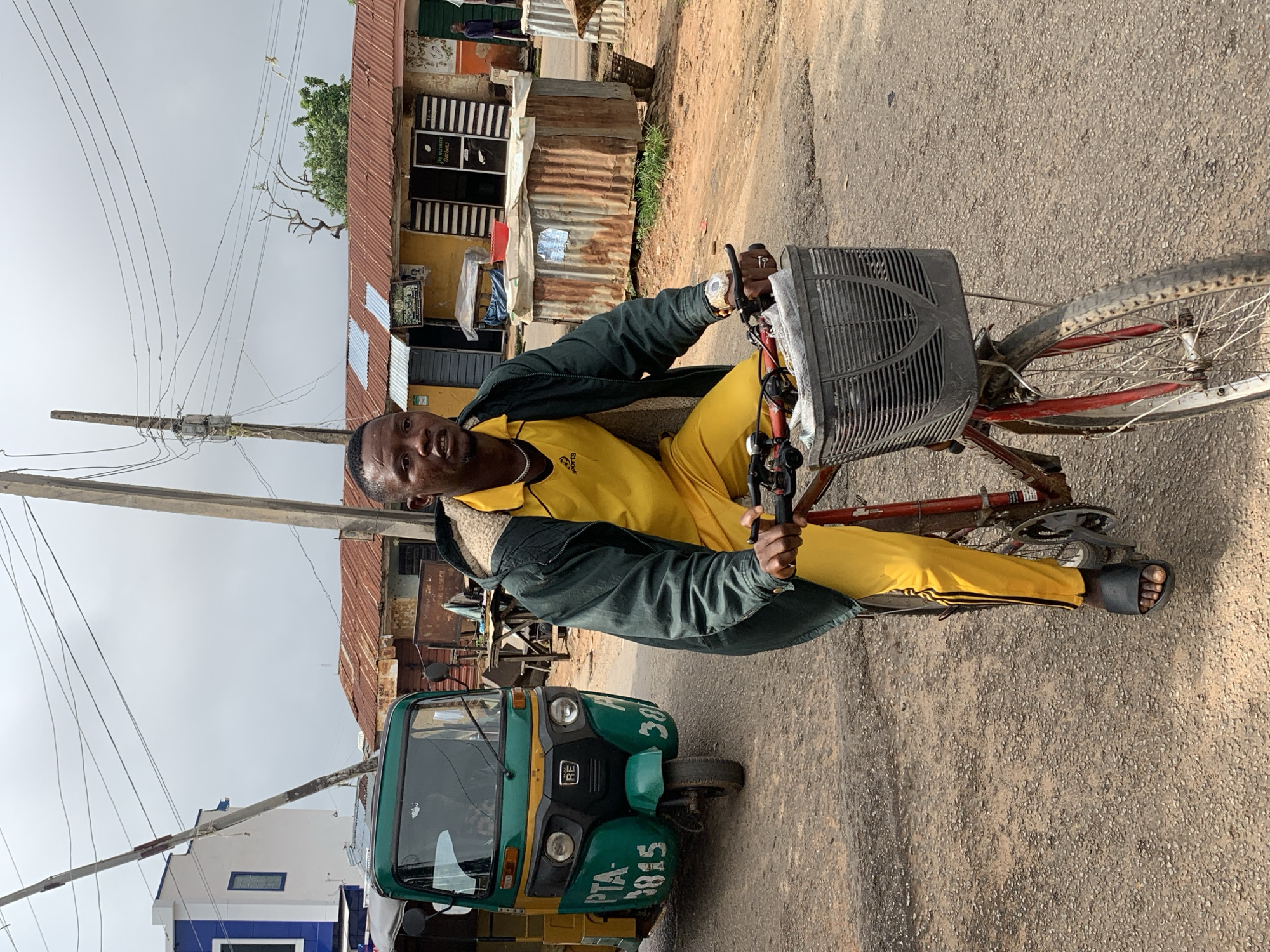 A man in a yellow tracksuit and dark jacket rides a bicycle with a basket in front. He is on a street in Jos, Nigeria, with buildings and a three-wheeled vehicle in the background. The sky is overcast.