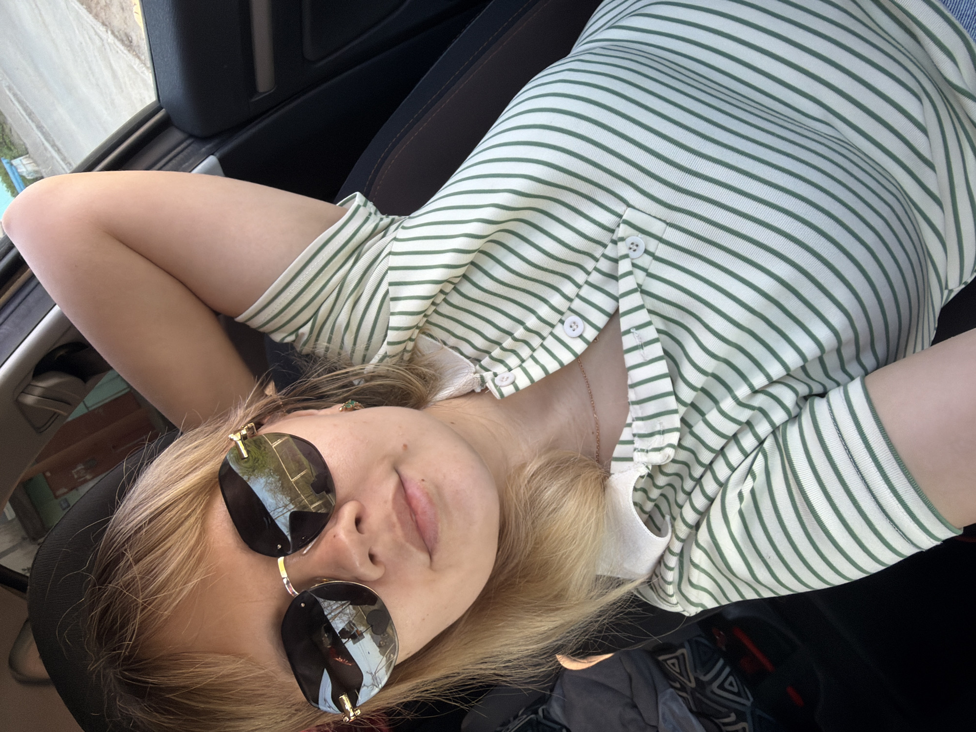 A person lies back in a car seat, wearing sunglasses and a striped shirt. The location is unknown, but possibly in the city of Chaikovsky, Russia. A car window is visible, along with car interior features. The person appears relaxed, with arms behind their head.