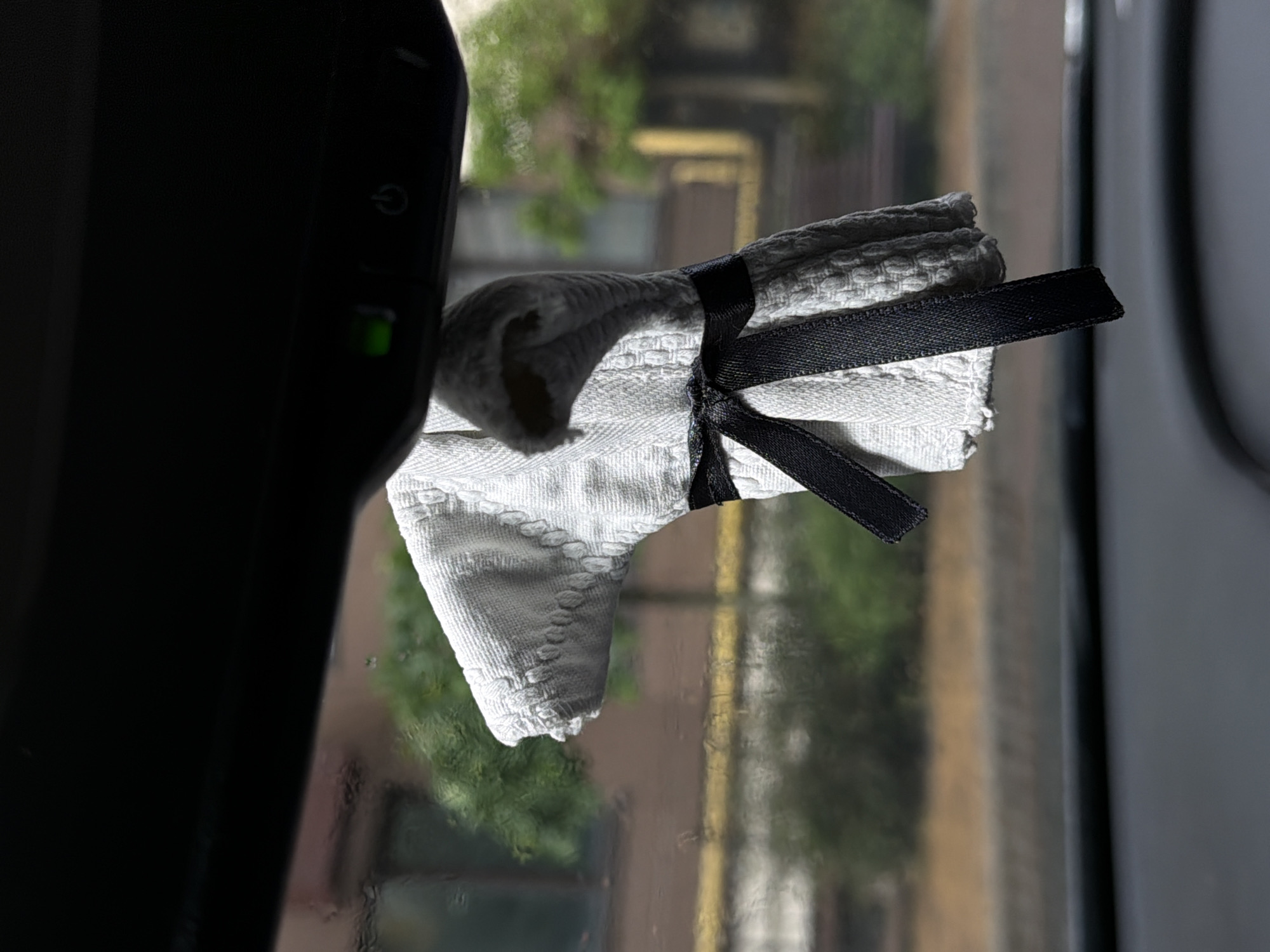 The image displays a close-up, slightly tilted view from an interior space, looking out a window in Sofiivska Borshchahivka, Ukraine. On the left side, a dark, angled electronic device is prominently featured, likely a dashcam or an integrated rearview mirror component. A small, illuminated green indicator light and a power symbol (a circle with a vertical line) are visible on its surface.
Adjacent to the device, a folded white cloth, appearing to be made of a textured or waffle-knit fabric, is neatly tied with a black satin ribbon. The ribbon forms a small knot, with its ends extending horizontally. The cloth seems to be positioned in the foreground, possibly resting on a surface or hanging.
The background, seen through the window, is significantly out of focus, indicating a shallow depth of field. This blurred outdoor scene reveals patches of vibrant green foliage, likely trees or bushes, and indistinct shapes of structures with brown or yellowish hues, suggesting distant buildings. The window surface itself appears to have smudges or moisture droplets, hinting at a potentially rainy or humid environment. The lighting suggests it is daytime, possibly under an overcast sky. No people are visible, and the scene is static, focused on the arrangement of the device and the tied cloth.