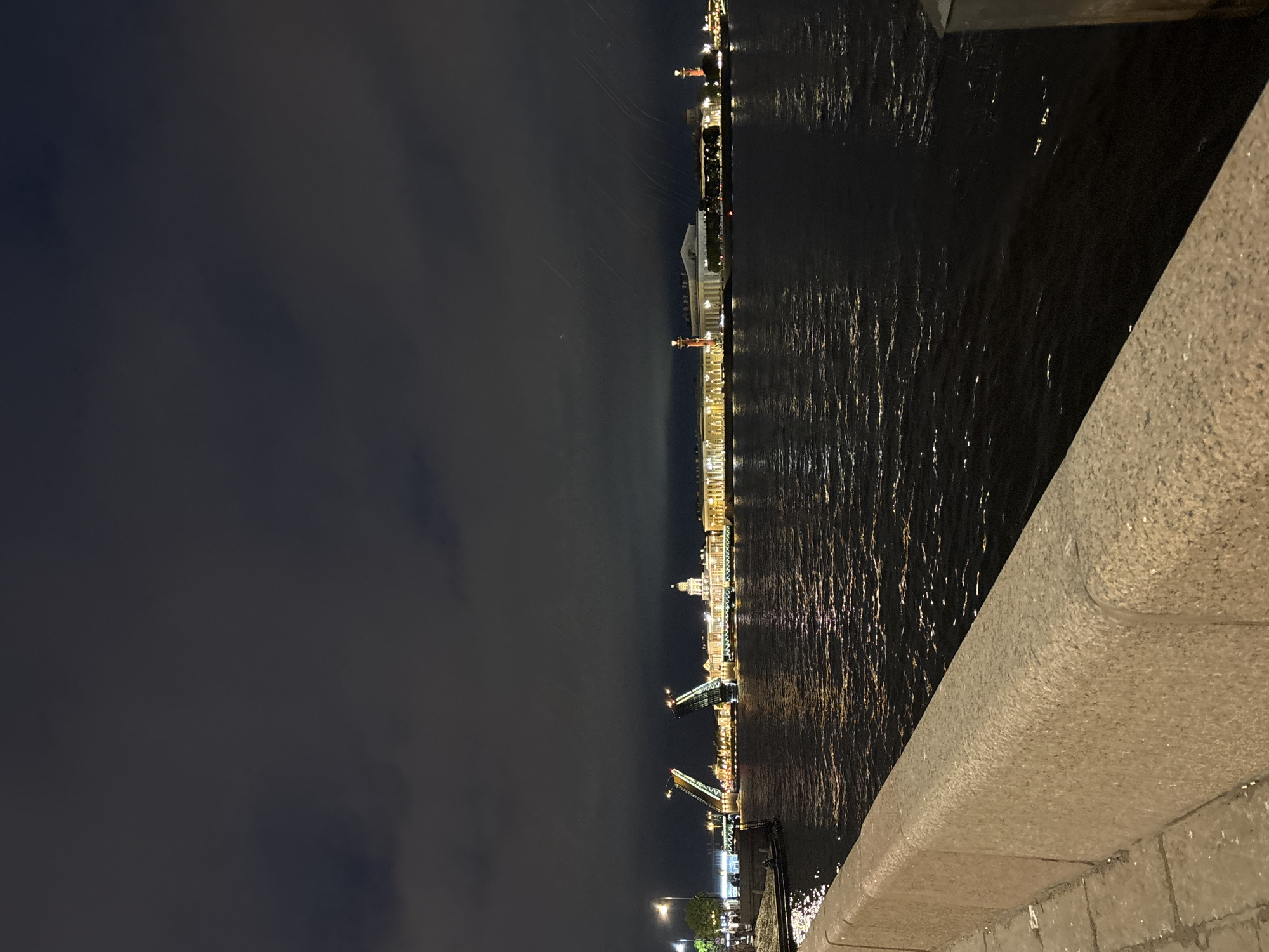 The image depicts a nighttime view of Saint Petersburg, Russia, from a bridge. The Palace Bridge is open, its halves raised, illuminated by lights. A river reflects lights from buildings along the embankment. Buildings include the Winter Palace, Admiralty, and Rostral Columns. The night sky is dark with clouds. A stone embankment edge is in the foreground.