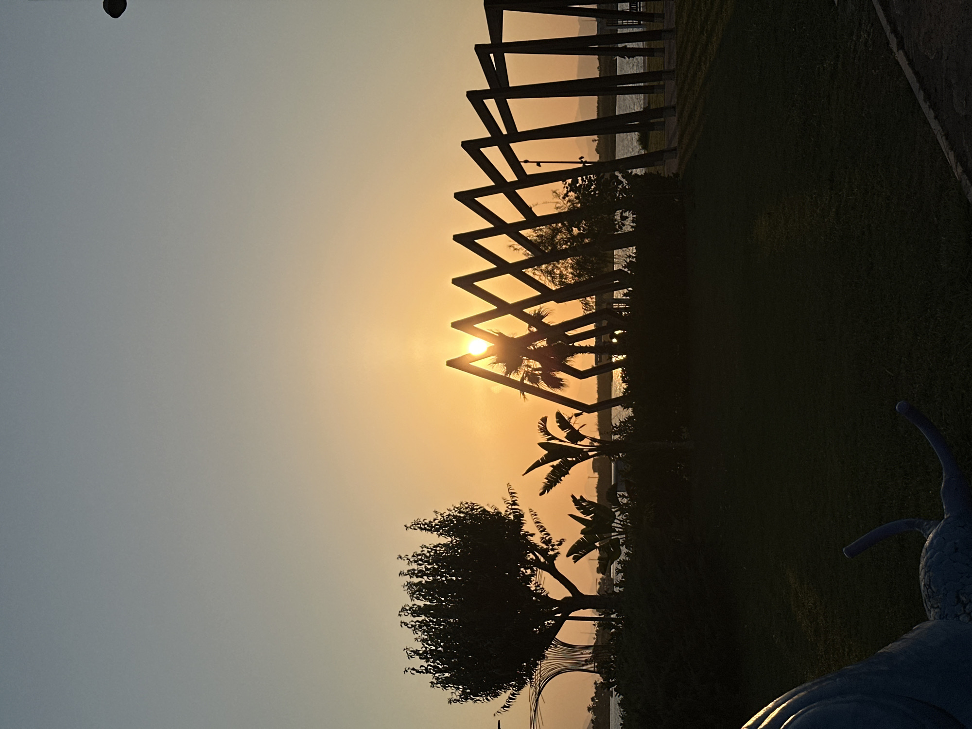 The image captures a sunset at a waterfront park in Fethiye, Turkey. The sun is setting behind a striking abstract wooden sculpture with a zigzag pattern, casting a warm, golden light across the sky. The sun's rays are visible peeking through the gaps in the sculpture, creating a dramatic effect. In the foreground, the silhouettes of trees and lush vegetation are prominent, with a body of water visible in the background, reflecting the sky's colors. To the left, a large, stylized sculpture resembling a blue snail is partially visible, suggesting a recreational or artistic area. The overall scene is peaceful, with the natural beauty of the sunset enhanced by the man-made structures.