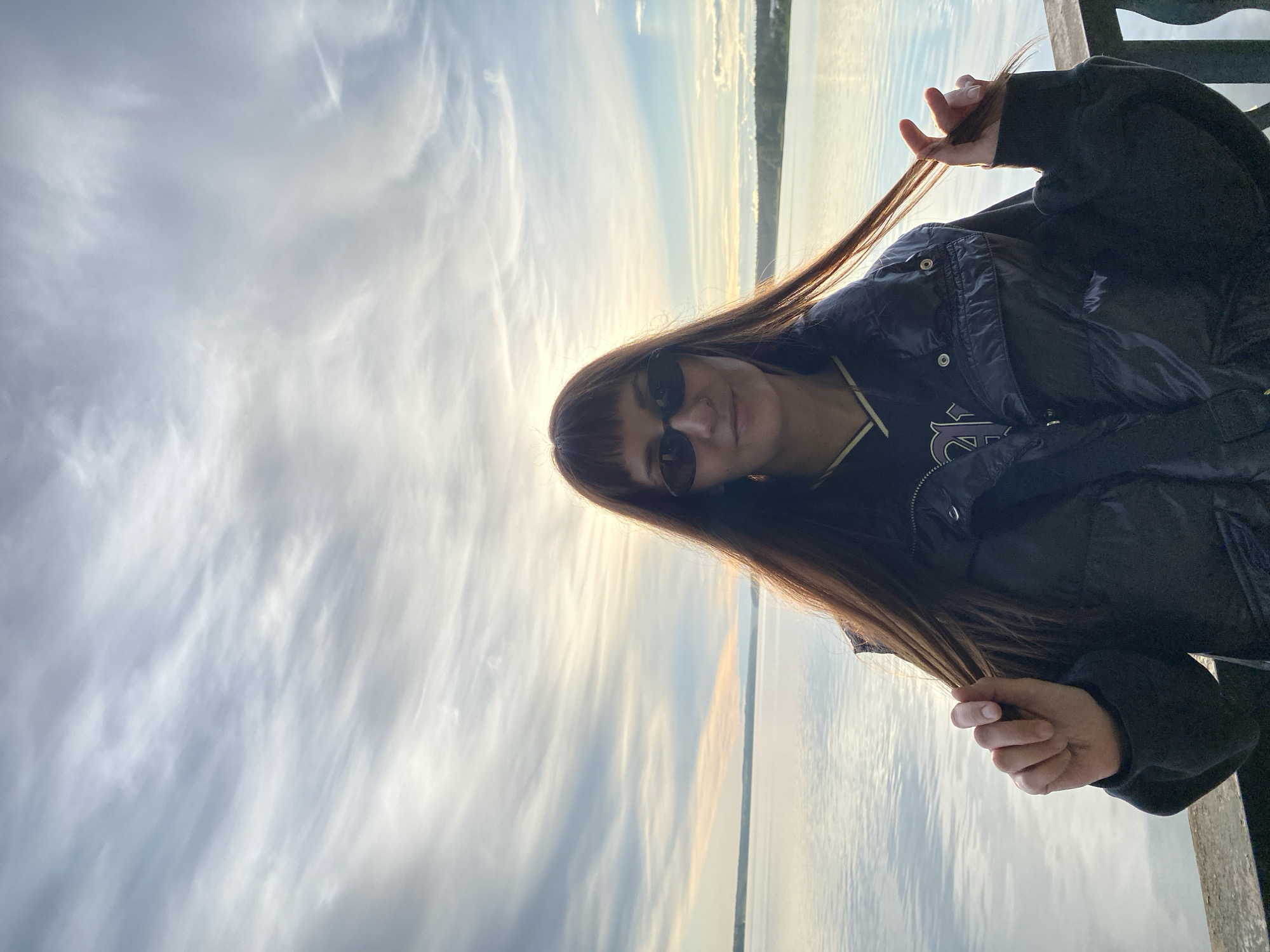 A woman with long brown hair and sunglasses is holding a strand of her hair in front of a cloudy sky and a body of water. She is wearing a black jacket. The location is likely near a body of water in Votkinsk, Russia, given the presence of water and the subject's likely connection to the city. There is no evidence in the image that confirms the specific location in Votkinsk.