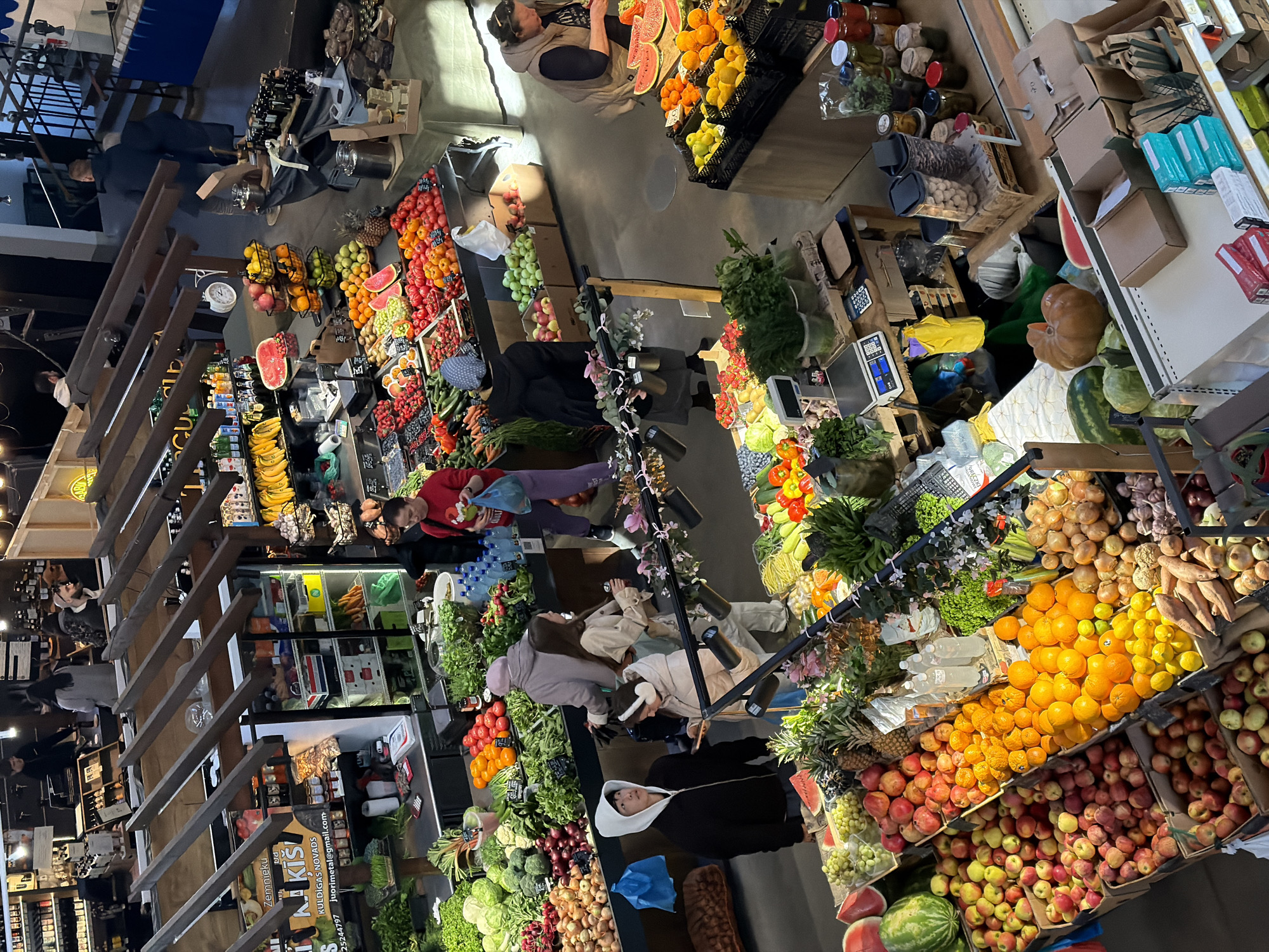 This media file captures a busy indoor market, likely a farmer's market or specialty grocery store, from an elevated perspective. The scene is filled with numerous stalls displaying a wide variety of fresh produce and other food items.
Several people are visible throughout the market, engaged in shopping and browsing. Individuals are seen selecting fruits and vegetables, standing near checkout areas, or moving between the different stalls. Some people appear to be market staff, tending to the displays or assisting customers. The activity suggests a vibrant commercial environment focused on fresh food.
The market is richly stocked with an extensive selection of fruits and vegetables. Prominent displays include large quantities of apples (various red, green, and yellow types), oranges, lemons, mandarins, bananas, watermelons, pineapples, grapes, pomegranates, strawberries, and tomatoes. Vegetables such as cucumbers, carrots, onions, garlic, various leafy greens, and squash or pumpkins are also abundantly showcased. In addition to fresh produce, there are shelves with packaged goods, including jars of preserves or pickled items, bottles of beverages, and bags of nuts or dried fruits. The products are neatly arranged in wooden crates, baskets, and on shelves.
Notable details include the market's interior architecture, featuring wooden beams and structures. Artificial overhead lighting and spotlights illuminate the displays, creating a bright and inviting atmosphere. A large wall clock is visible in one section, indicating the time is approximately 1:55. Visible text on a sign includes "Zemnieku" (Latvian for "farmer's") and "KULDĪGAS NOVADS" (Kuldīga Municipality), confirming the location context of Riga, Latvia. The market appears well-organized and maintained, presenting an array of fresh, high-quality goods.