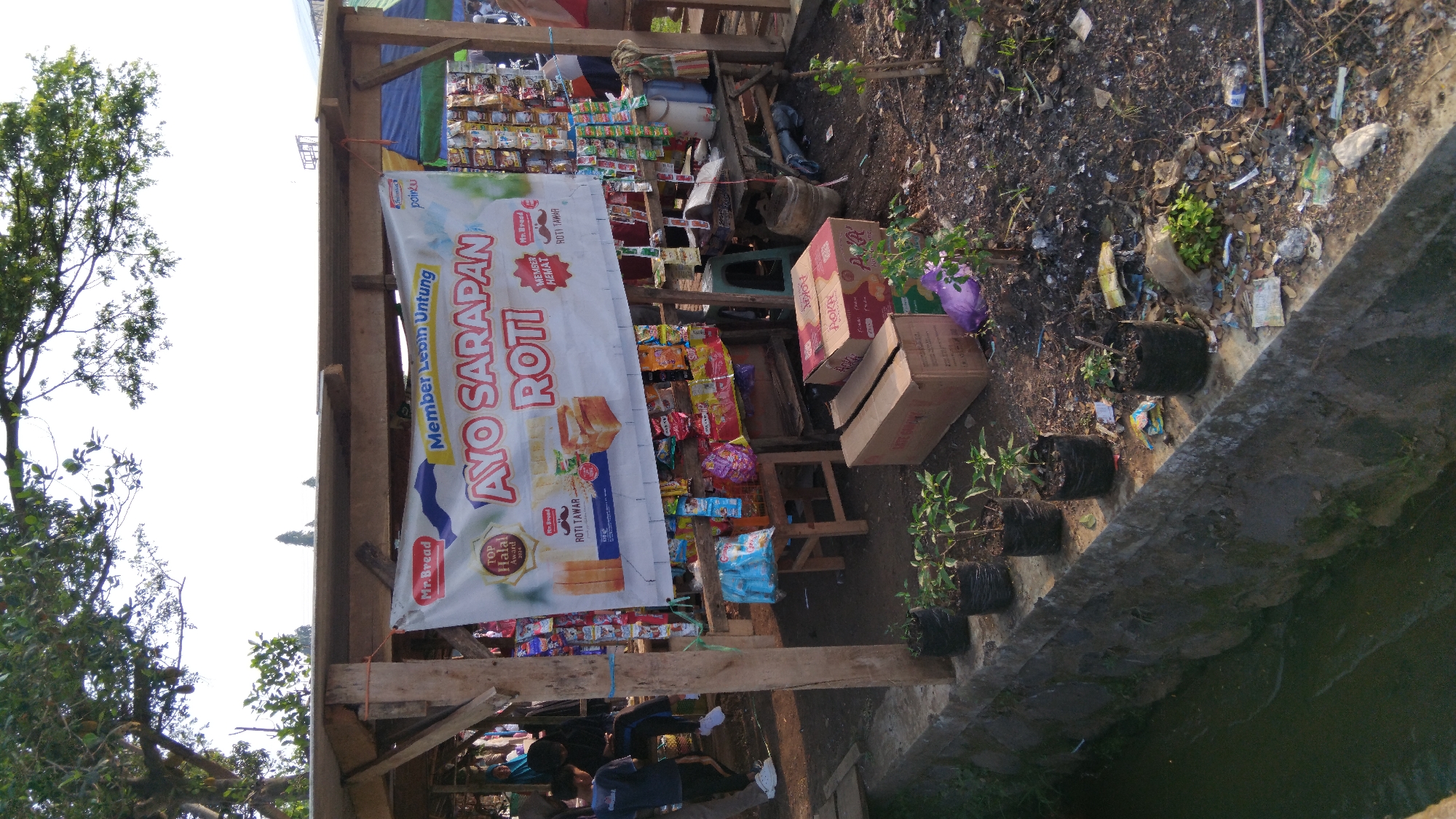The image shows a small roadside stall in Sembojo, Indonesia, selling bread and snacks. A banner on the stall reads "AYO SARAPAN ROTI" (Let's have bread for breakfast). Several people are visible in the background, some of whom appear to be customers or vendors. The stall is stocked with various food items, and there are also some plants in pots in the foreground. The environment appears to be a somewhat unkempt area, with some debris visible on the ground. The overall impression is of a humble, everyday scene in a local Indonesian community.