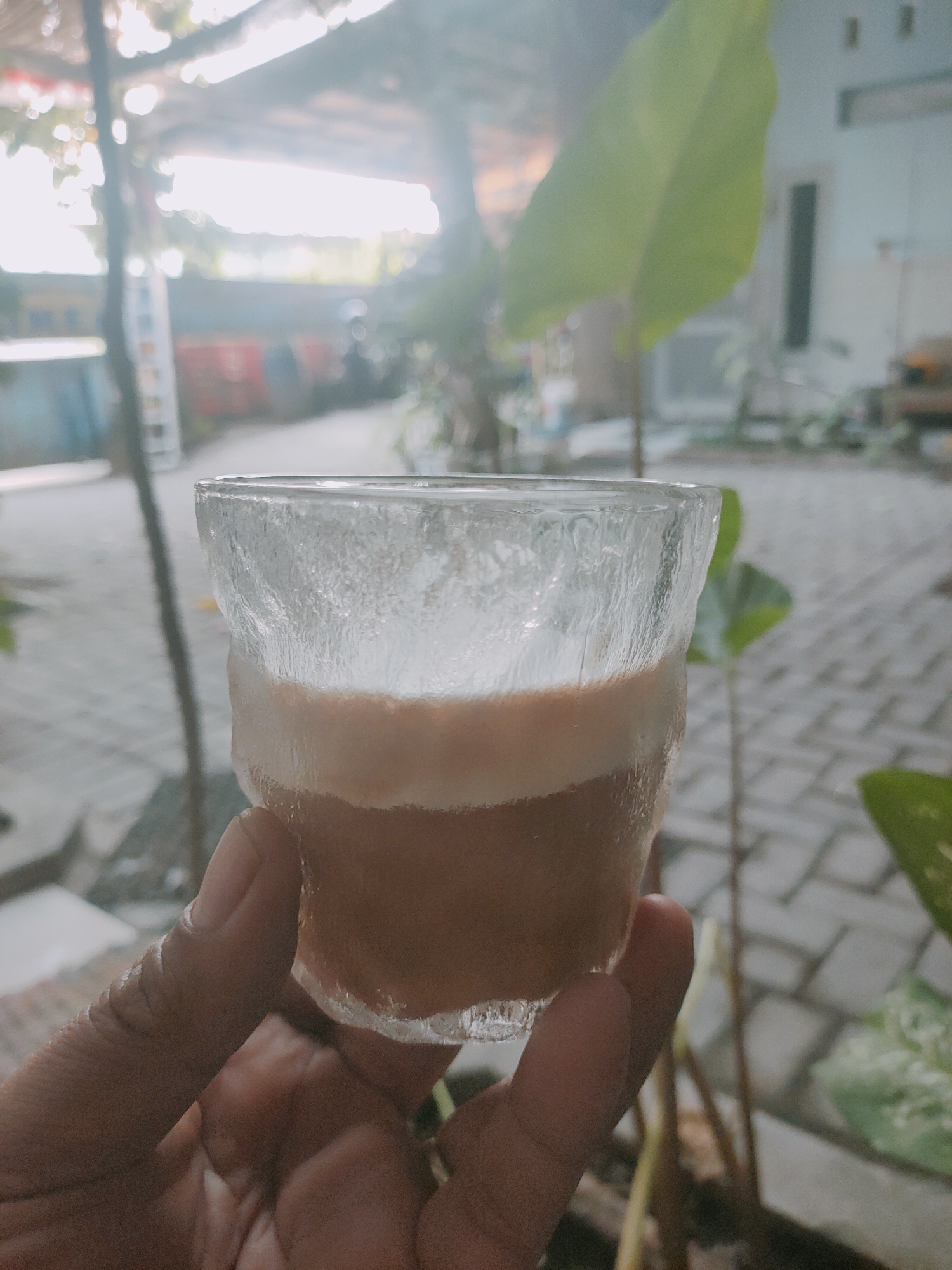 A hand is holding a glass of iced coffee. The glass is made of textured glass, and condensation can be seen on the outside. The iced coffee is layered, with a lighter colored foam on top and a darker coffee at the bottom. The background is blurred but appears to be an outdoor setting, possibly a patio or garden, with a paved walkway and some greenery. The scene suggests a moment of relaxation or enjoyment of a beverage in a warm climate.
