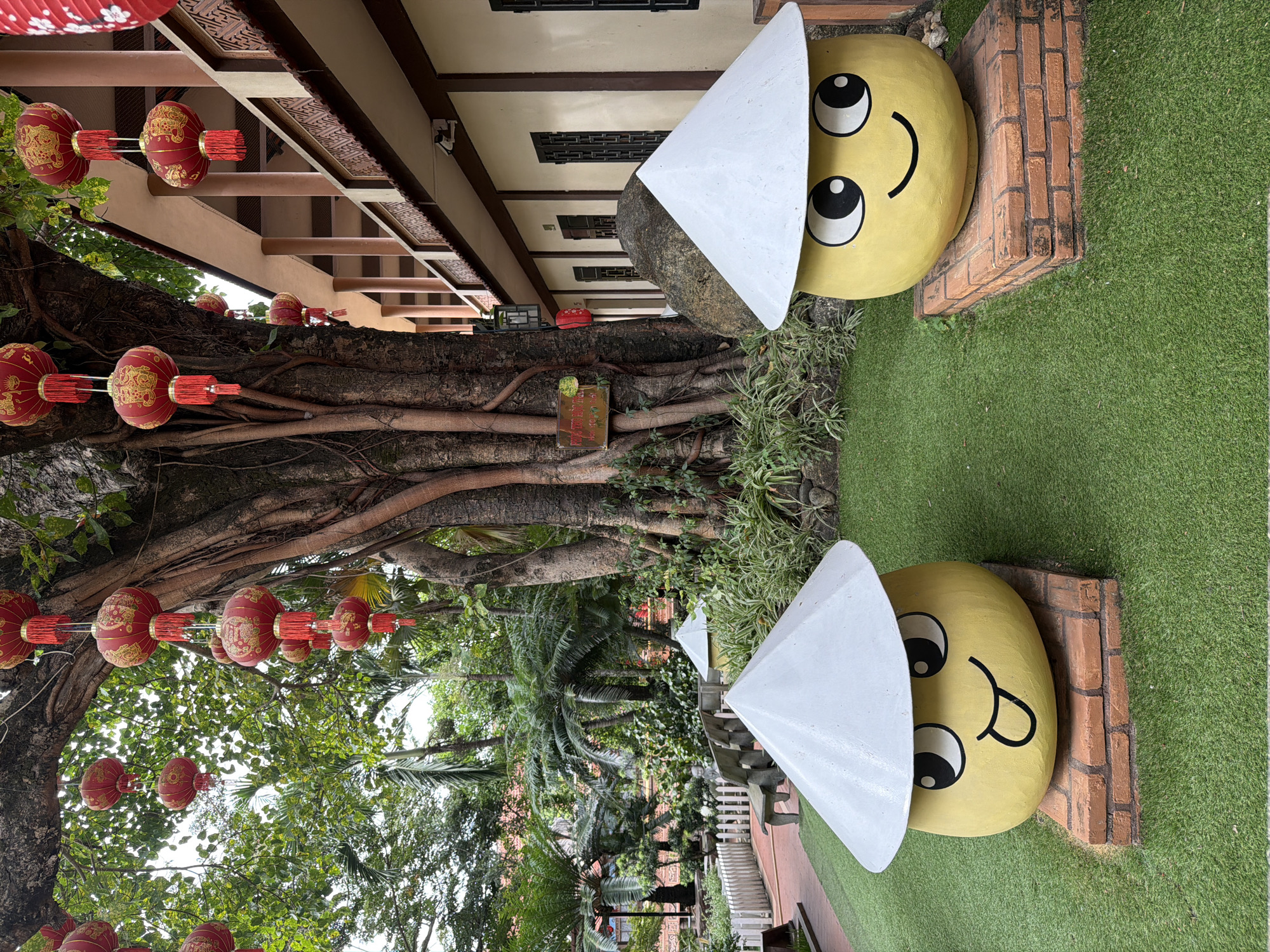 Two yellow, spherical sculptures with large black and white eyes and smiling mouths are situated on a bright green, artificial lawn. They are each topped with a conical white hat. The sculpture on the left is partially obscured by the sculpture on the right. Both sculptures are elevated on a rectangular base made of red bricks. In the background, a large tree with exposed roots dominates the scene. Red lanterns are strung from the tree's branches and from a nearby building. The building is a light tan color with a covered walkway supported by brown pillars. A stone sits behind the right sculpture. The environment appears to be a garden with lush green foliage and palm trees. The scene is likely located in Hoi An, Vietnam, as suggested by the presence of the conical hats and the overall architectural style.