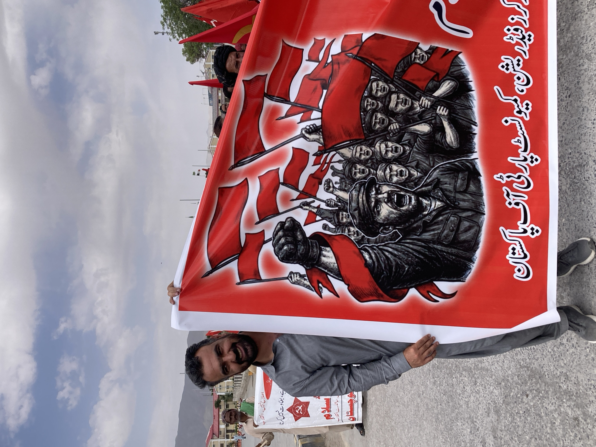 Here's a concise description of the image:
The image shows a man holding a large red banner with a black and white illustration of a crowd of people holding red flags. The banner also has text in Urdu script on the right side. The man holding the banner is smiling. Behind him, another banner is partially visible, also containing text in Urdu. Additional people are in the background. The location appears to be an outdoor area in Quetta City Tehsil, Pakistan, based on the visible architecture and the text on the banners.