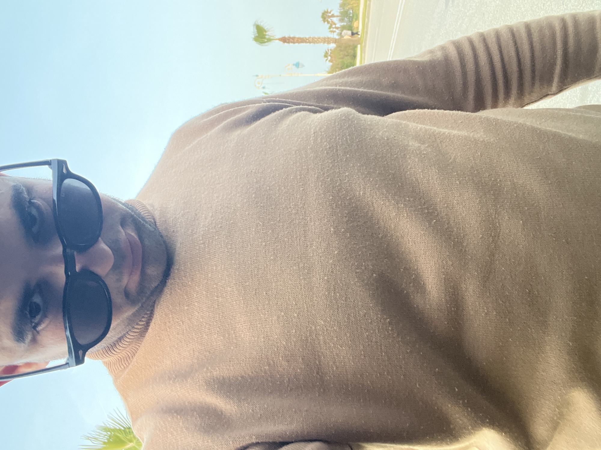 The image captures a close-up, low-angle perspective of a person, likely a selfie. The subject, visible from the chest up, is wearing dark sunglasses that cover their eyes, and a light brown or tan long-sleeved top, possibly a sweater, which shows visible pilling on its fabric. Some facial hair, appearing as stubble or a short beard, is visible around the chin and cheeks.
The background, predominantly a clear, bright blue sky, indicates a sunny daytime setting. In the distance, several palm trees are visible, along with what appears to be a paved road or pathway featuring white line markings. Decorative streetlights or poles can also be seen in the background. The overall scene suggests an outdoor environment in a warm climate, consistent with the provided location context of Martil, Morocco. There is no visible text in the image.