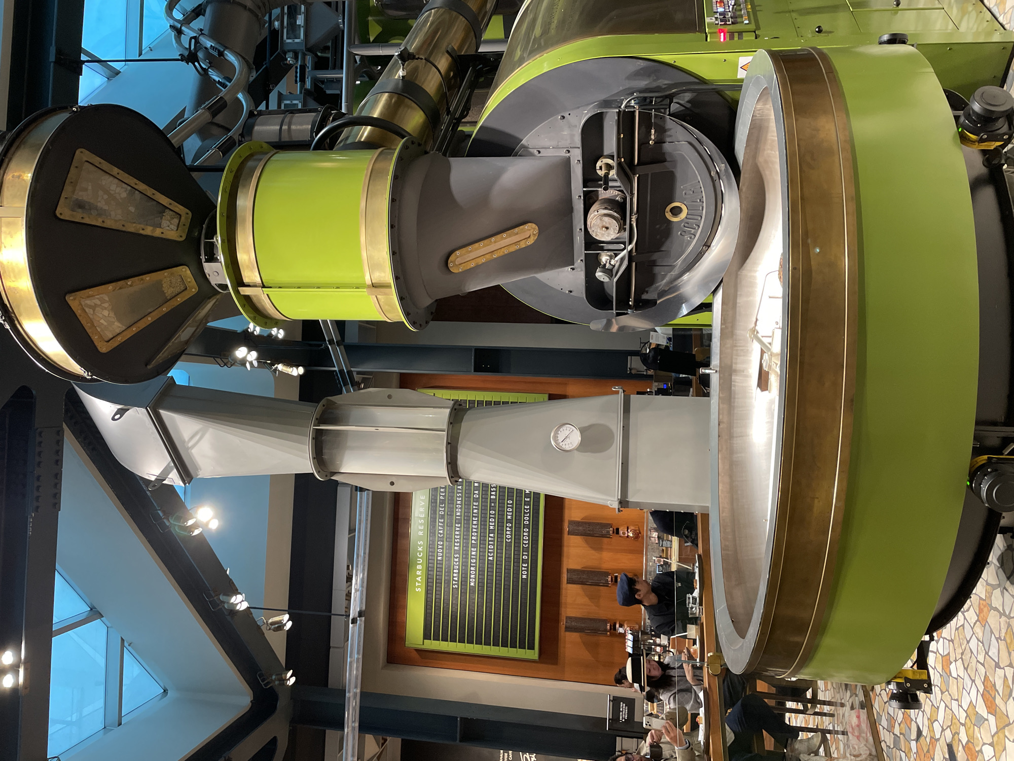 The image shows the interior of the Starbucks Reserve Roastery in Milan, Italy. Prominent is a large, industrial coffee roasting machine in green, gray, and brass colors. Sections of the machine include a large cylinder and ducts. To the left of the roaster, a menu board displays "Starbucks Reserve" and lists various coffee types and flavor profiles. People can be seen at a counter, some seated and looking at their phones while others are standing. The architecture features skylights and a modern design.