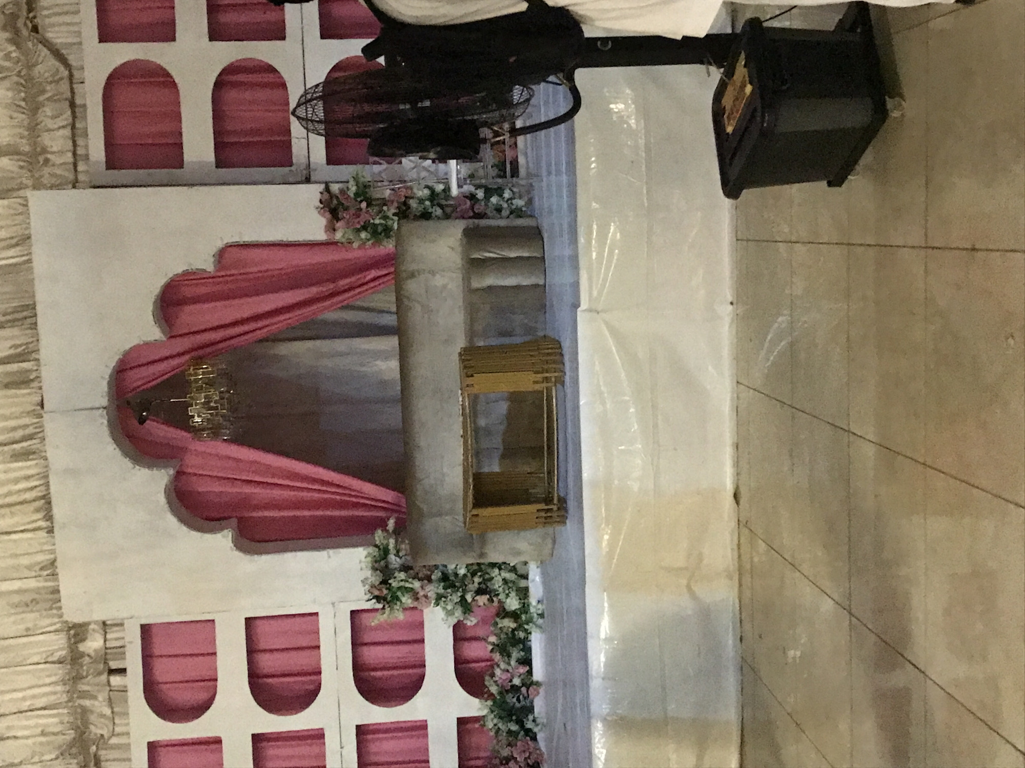 The image depicts a stage setup, possibly for an event, likely indoors. The stage backdrop features a white wall with pink arched details. A pink draped fabric adorns the center, and floral arrangements sit at the base. A gray cylindrical structure stands on the stage, with a small wooden crate nearby. A fan and a black box are positioned above, potentially for lighting or sound. The stage floor is tiled and the image was taken in the city of Oko Erin, Nigeria.