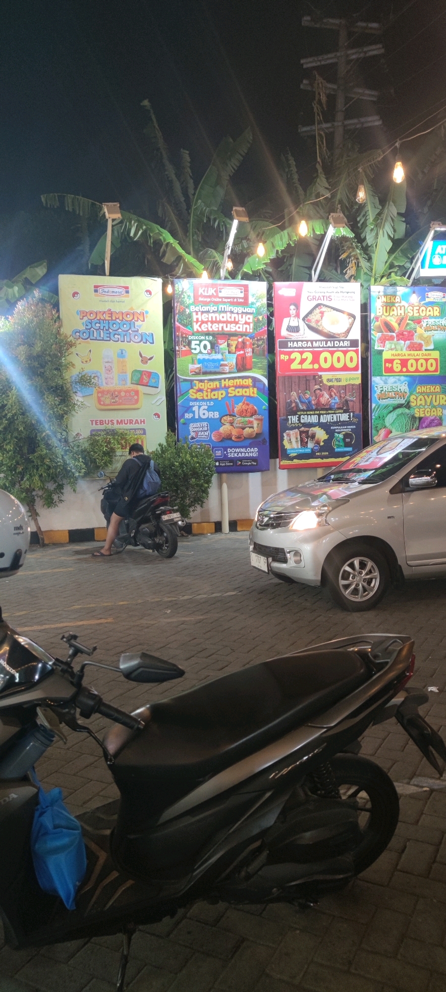 This image depicts an outdoor scene at night, likely a street or parking area, with several large advertisements displayed vertically. The advertisements feature vibrant graphics and text, promoting products and offers from brands such as Indomaret and KUK. One banner advertises a "Pokémon School Collection," while another promotes "Belanja Minatnya Hematnya Keterusan!" with discounts and food imagery. A third banner, partially visible, shows "Aneka Buah Segar" and "Aneka Sayur Segar."
In the foreground, two motorcycles are parked. One is a dark-colored scooter with a blue bag hanging from its left side. The other motorcycle is mostly out of frame but shows a portion of its handlebars and seat.
A silver car is parked in the middle ground, with its headlights illuminated. A person is seated on the dark-colored scooter in the middle ground, facing away from the camera, wearing a dark shirt and shorts, with a backpack slung over their shoulders.
The background features lush green foliage, likely banana trees, and decorative string lights that contribute to the nighttime ambiance. The ground is paved with interlocking bricks.
While the specific location of "Keboansikep, Indonesia" is mentioned, there are no visual cues within the image itself that definitively confirm this location. However, the advertisements are in Indonesian, suggesting an Indonesian setting.
