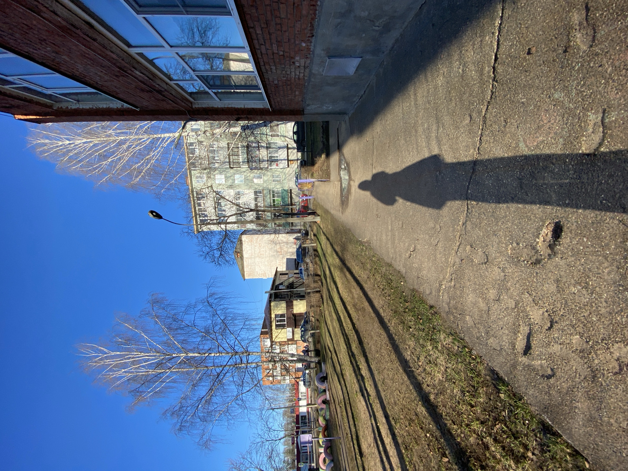 The photograph captures a street scene in Noviy, Russia. The image is taken at ground level, angled upwards. On the left, a brick building with multi-pane windows reflects the clear blue sky. A bare tree stands in front. To the right is a concrete walkway, casting a shadow of a person with a hat, with bare feet. Beyond the walkway, a grassy area leads to a playground with tires laid out. Several apartment buildings stand in the background. A lamppost is visible near the buildings. Cars are also parked in the background, and the light suggests it is daytime.