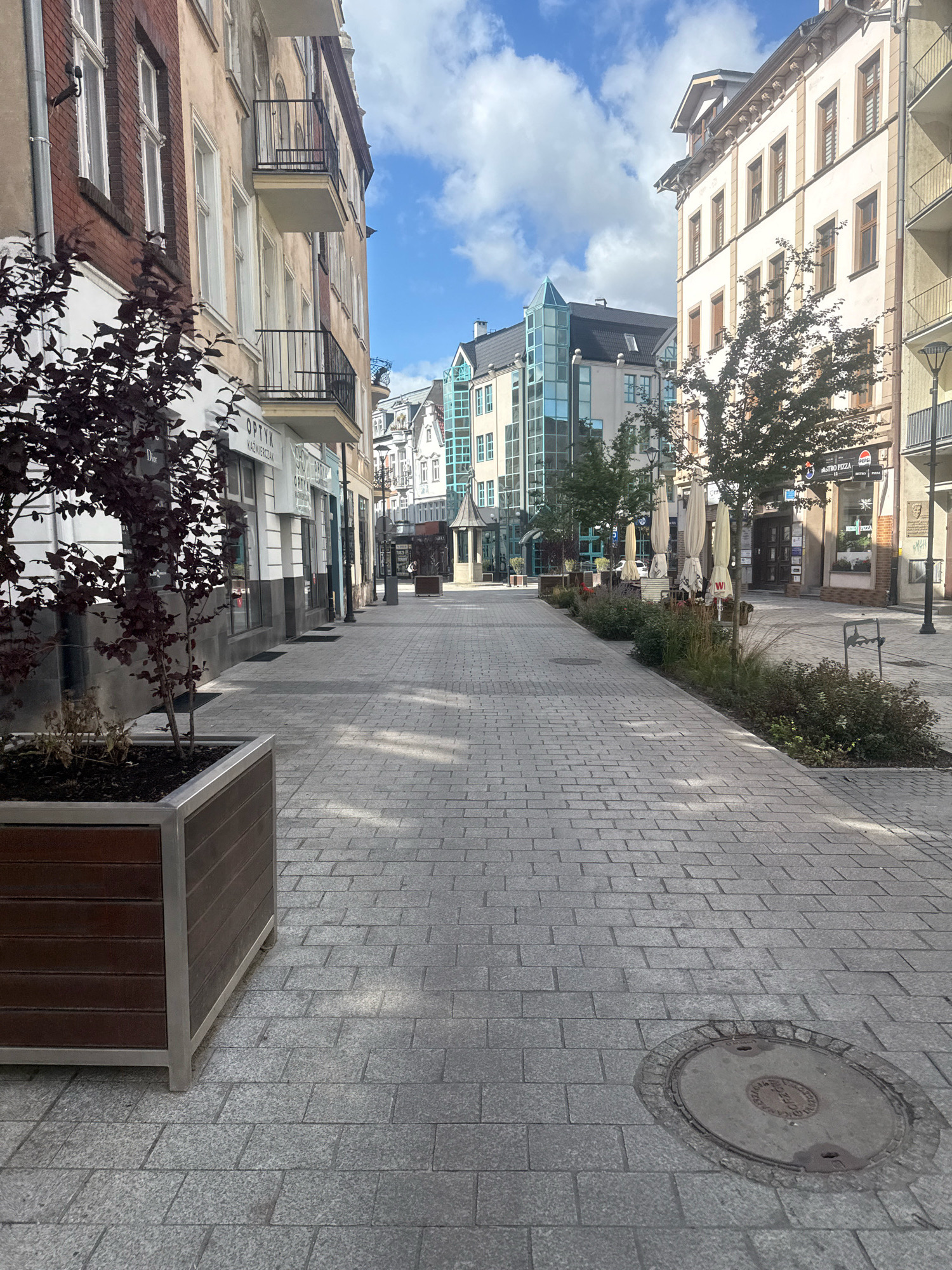 This is a street scene in Gorzow, Poland, with buildings lining both sides of a pedestrian walkway paved with grey stone bricks. On the left, a building with red brick lower floors and beige upper floors features balconies with black railings. A large wooden planter with a dark-leaved tree is positioned in the foreground. Shops with large glass windows are visible along this side of the street, with signage including "OPTYK KAZMIERCZAN". On the right, a lighter colored building has multiple floors and windows, with balconies. Further down the street, there are trees, outdoor umbrellas, and the entrance to a "BISTRO PIZZA". The sky is blue with scattered white clouds. A circular manhole cover is visible in the pavement on the right side of the walkway. The scene appears quiet, with no visible people.