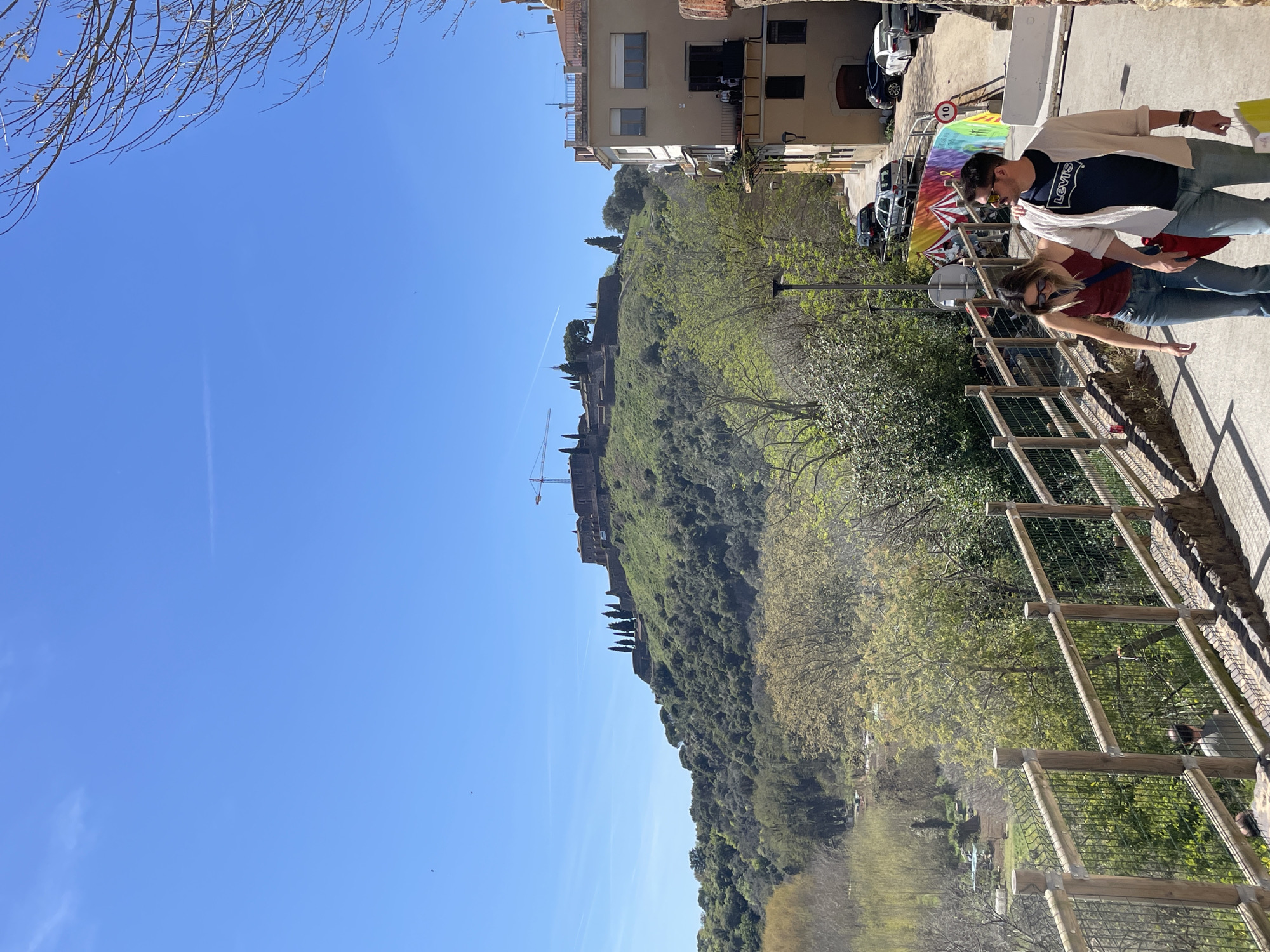 Captured in Hostalric, Spain, the image presents an outdoor scene under a clear blue sky. A green, tree-covered hill dominates the view, topped by a castle with ongoing construction indicated by a crane. In the foreground, a couple walks along a fenced walkway, their attire casual. Cars are parked nearby, and buildings stand adjacent to the walkway. The image is taken from a slightly elevated perspective, overlooking the slope.