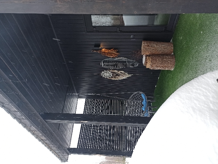 The image displays a covered outdoor patio area of a structure located in Nizhneye Zadolgoye, Russia, under conditions of light snowfall. The building facade is constructed of dark, vertical wooden siding, with a corresponding dark wooden overhead roof structure supported by two prominent dark vertical beams. The ground immediately beyond the patio is covered in a substantial layer of white snow, forming a rounded mound in the lower-left foreground. Individual snowflakes are observable against the dark building and roof surfaces.
The patio floor consists of artificial green turf. A dark, egg-shaped hanging chair, fitted with a blue cushion, is suspended by a chain from the overhead structure between the two support beams. Adorning the dark building wall are two distinct decorative macrame or woven wall hangings, predominantly light in color, situated to the left of a bundle of dried branches, possibly a venik. Beneath these wall decorations, two natural tree stump sections of varying heights stand vertically on the green turf. A dark, wall-mounted light fixture is positioned between the decorative items and a dark-framed glass door leading into the building. A dark fence-like structure with horizontal slats is visible behind the left support beam. The background reveals an overcast sky and faint silhouettes of bare trees. No people are present in the scene.