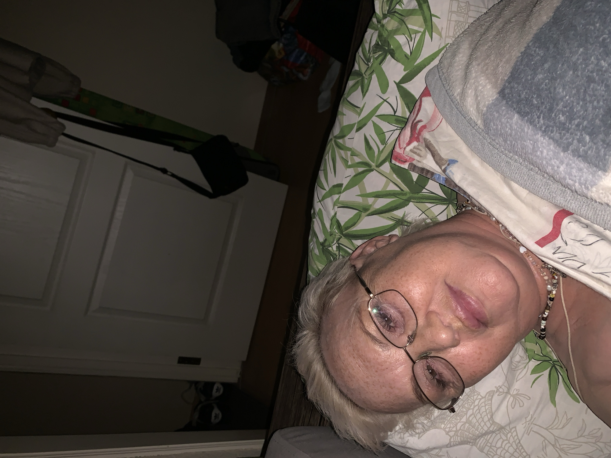 An elderly woman with blonde hair and eyeglasses is lying in bed, covered partially by a patterned blanket and a blue and white fuzzy blanket. The bedsheets have a green and white plant pattern. She is wearing a beaded necklace. The room is dimly lit. In the background is a white door with a black purse hanging on it. The location is likely inside a private residence, but there is no information to suggest the specific city of Chaikovsky, Russia.