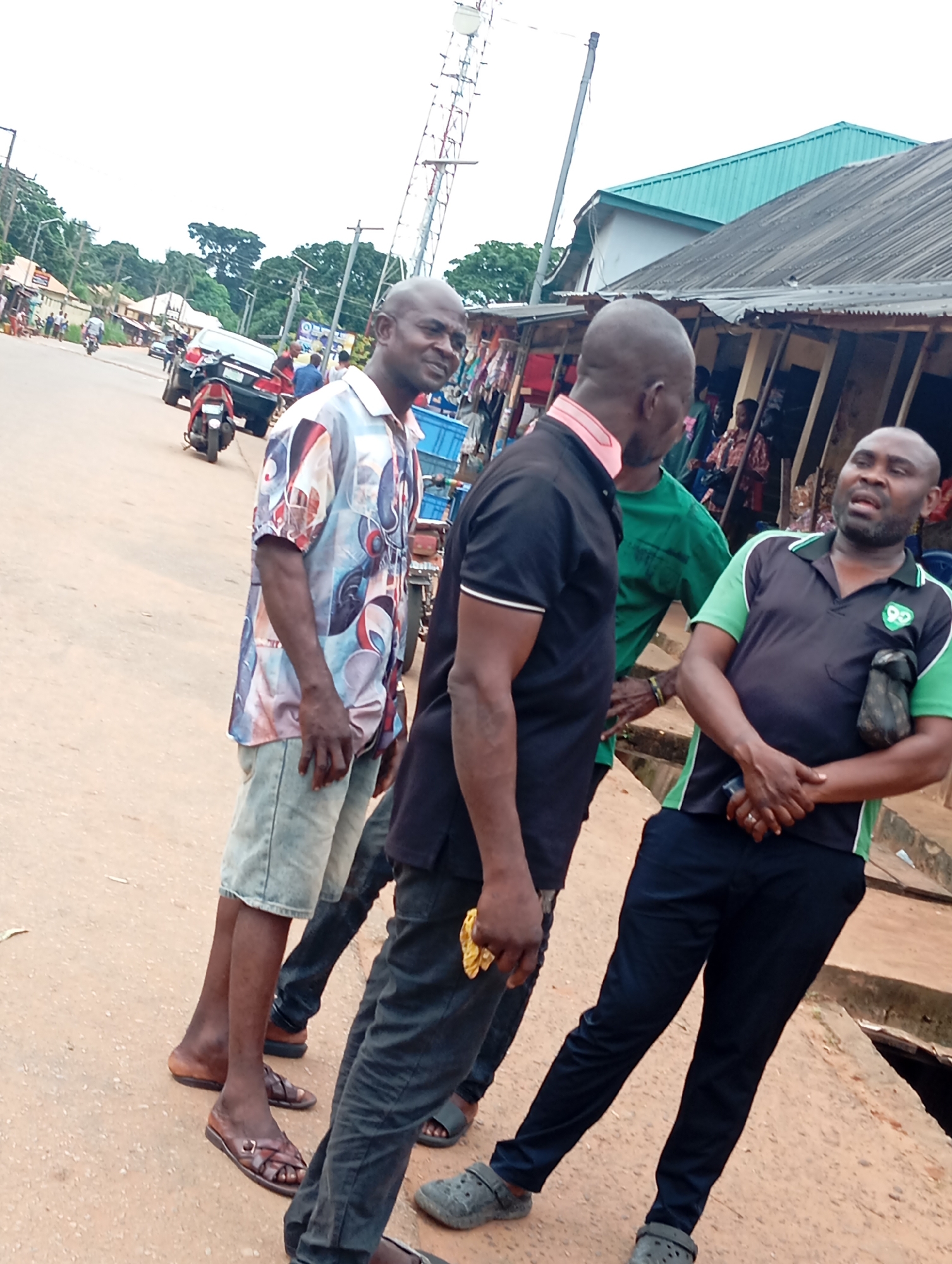 The image depicts an outdoor scene on a dusty, paved road in what is identified as Nsugpe, Nigeria. Three adult males are the primary subjects, positioned centrally in the frame. The man on the left wears a short-sleeved, colorful patterned shirt and light-colored denim shorts, standing with his legs apart and his right hand resting on his hip. To his right stands another man in a black polo shirt with a pink collar, his arms slightly bent. To the right of the man in black, a third man is visible from the chest down, wearing a dark green and black polo shirt with a logo on the left chest and dark trousers. He is speaking, with his mouth open and his hands clasped in front of him.
In the background, a busy street scene unfolds. Several motorcycles and a car are parked or in motion. Buildings line the right side of the road, some appearing to be shops or market stalls with corrugated metal roofs. Trees and utility poles are visible in the distance, suggesting a somewhat developed but potentially informal urban environment. The sky is overcast, indicating potentially diffused lighting conditions. The overall impression is a moment captured in a public space, possibly a market area or a thoroughfare in Nsugpe.