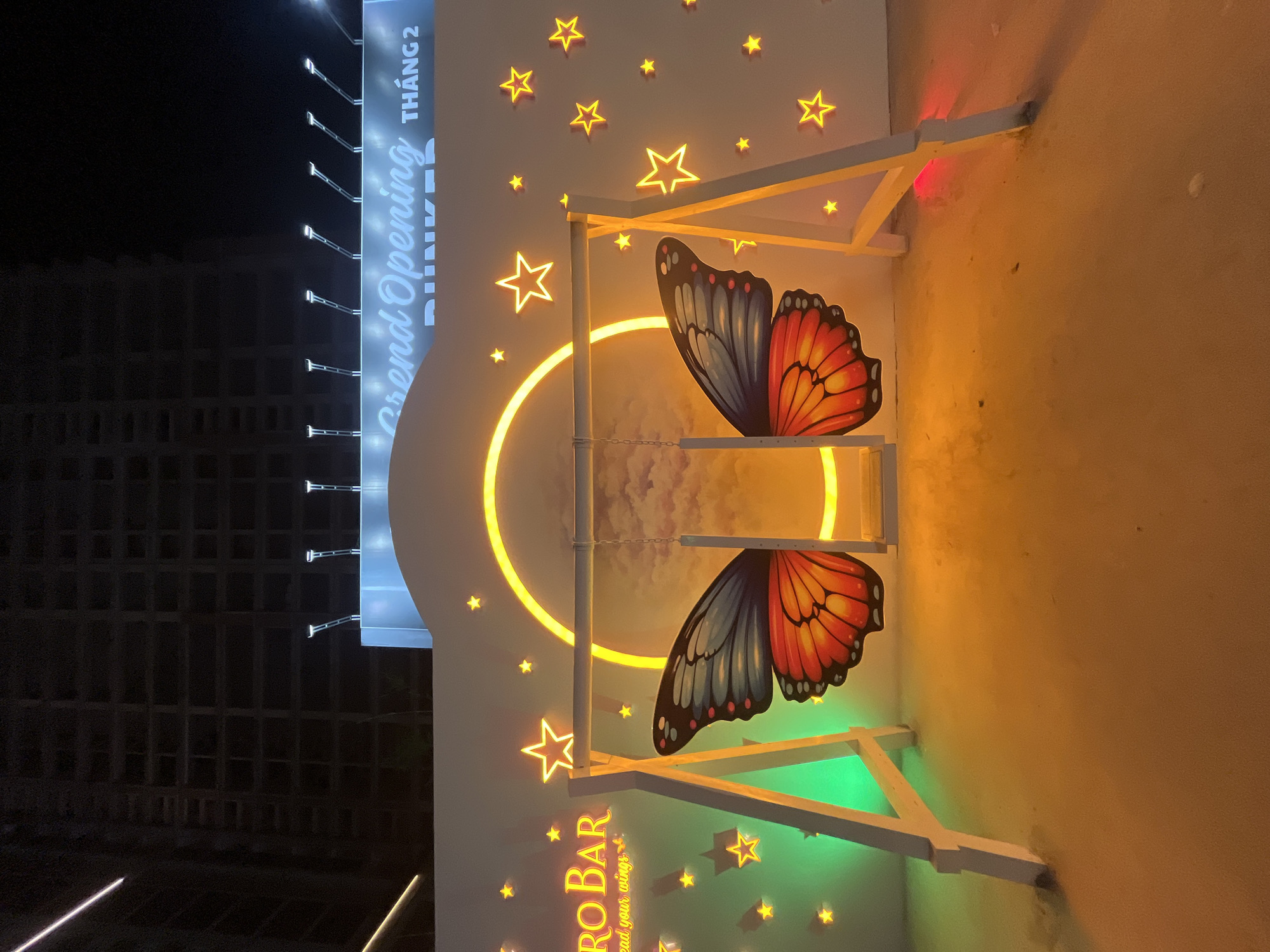 The image shows an outdoor nighttime scene in Da Nang, Vietnam, featuring a decorative swing adorned with butterfly wings. The swing is positioned in front of a white wall illuminated by neon lights. The wall displays various glowing stars and a large, circular neon light that resembles a moon. To the left of the swing, a neon sign reads "RO BAR" with the tagline "spread your wings." Above the wall, a large billboard with "Grand Opening THÁNG 2 BUNKER" is visible, along with the silhouette of a dark, unfinished high-rise building. The ground is sandy and illuminated by the neon lights, casting orange and green glows. No people are present in the image.