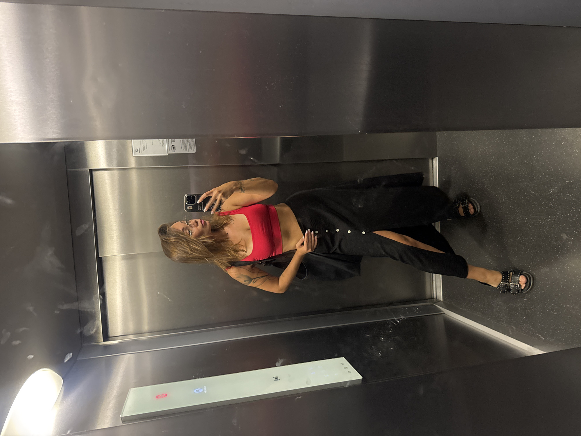 A woman is taking a mirror selfie inside an elevator. She is wearing a red crop top and a black maxi skirt with a slit. She has on black sandals with buckles and studs. A black tote bag is slung over her left shoulder. The elevator has stainless steel walls and a digital display panel on the left wall showing a red button at the top and other floor buttons below. The floor is dark gray with white speckles. The overall scene is utilitarian and modern, characteristic of an elevator found in a building, possibly a hotel or residential complex, in a city like Budva, Montenegro.