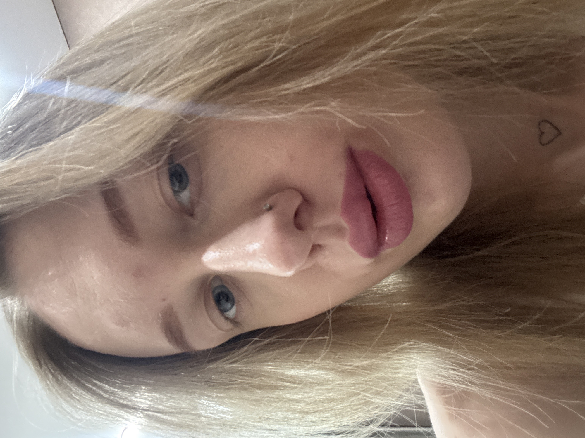 A close-up portrait shows a woman with fair skin, blonde hair, and blue eyes. She is tilted to the side, looking at the camera. Her lips are full and appear to be coated with lipstick. She has a nose piercing and a small heart-shaped tattoo is visible on her neck. The background is not visible, suggesting the image is tightly cropped. There is no information provided in the image to suggest the location of Ovoshchnoy, Russia or any city location.