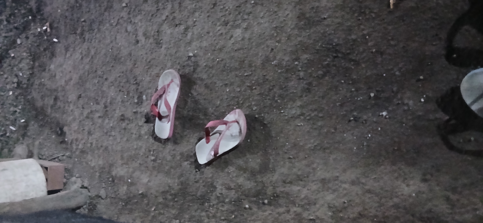 In Demangan, Indonesia, a ground-level view captures two pairs of white flip-flops with pink straps lying on a dirt and gravel surface. The scene suggests an outdoor environment, possibly a pathway or yard. The flip-flops appear worn and used. The lighting is dim, indicating it could be dusk or nighttime. There is a blurry object in the foreground, possibly a person's feet or shoes, casting a shadow.