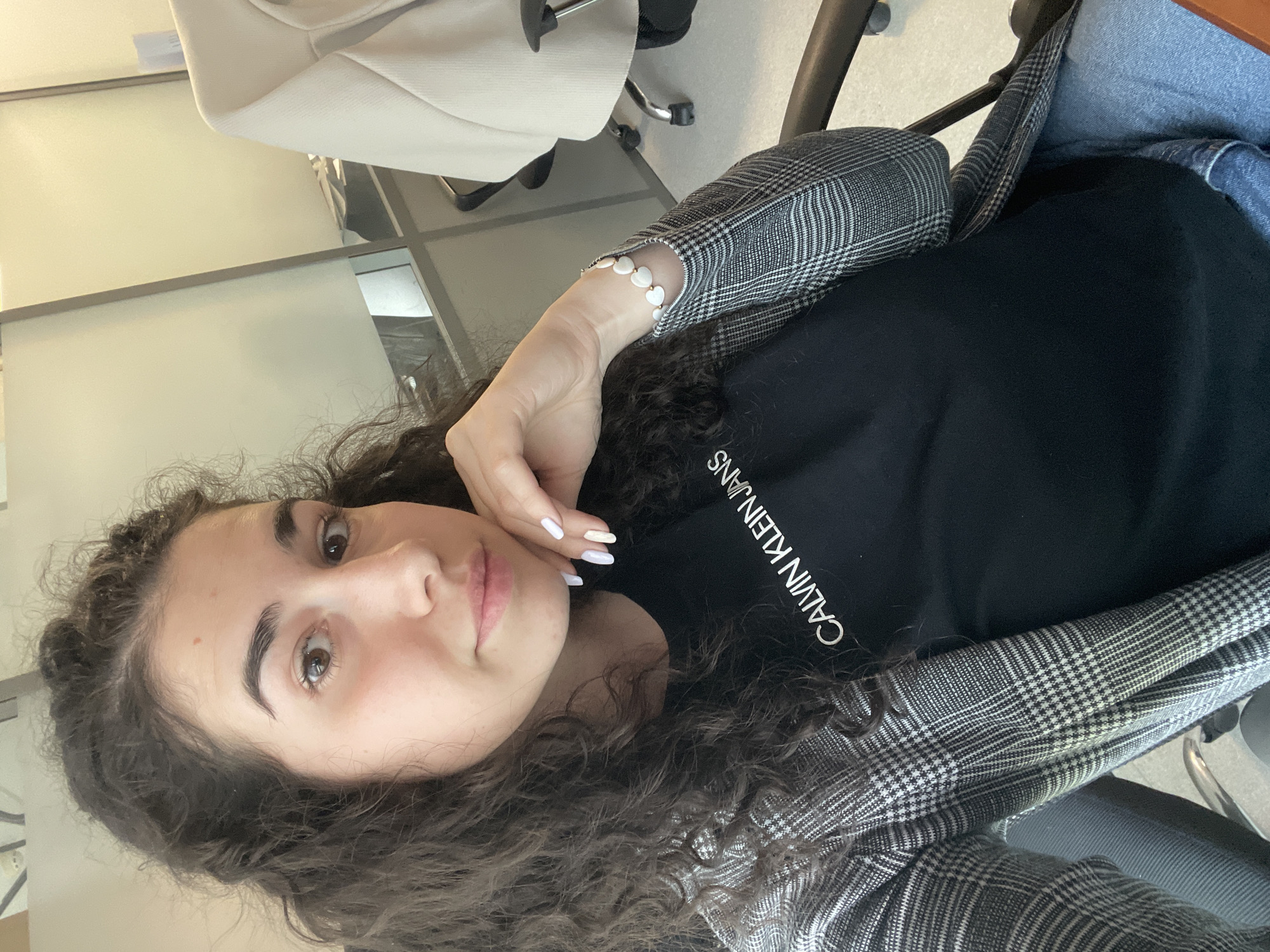 A young woman with curly dark hair and light skin is seated in an office chair, wearing a black Calvin Klein t-shirt and a grey plaid blazer. Her left hand, adorned with a pearl bracelet, rests near her face. The scene appears to be indoors, possibly in an office setting with white walls and office furniture. The location is indeterminate, and cannot be determined to be Sofia, Bulgaria, from the image alone.