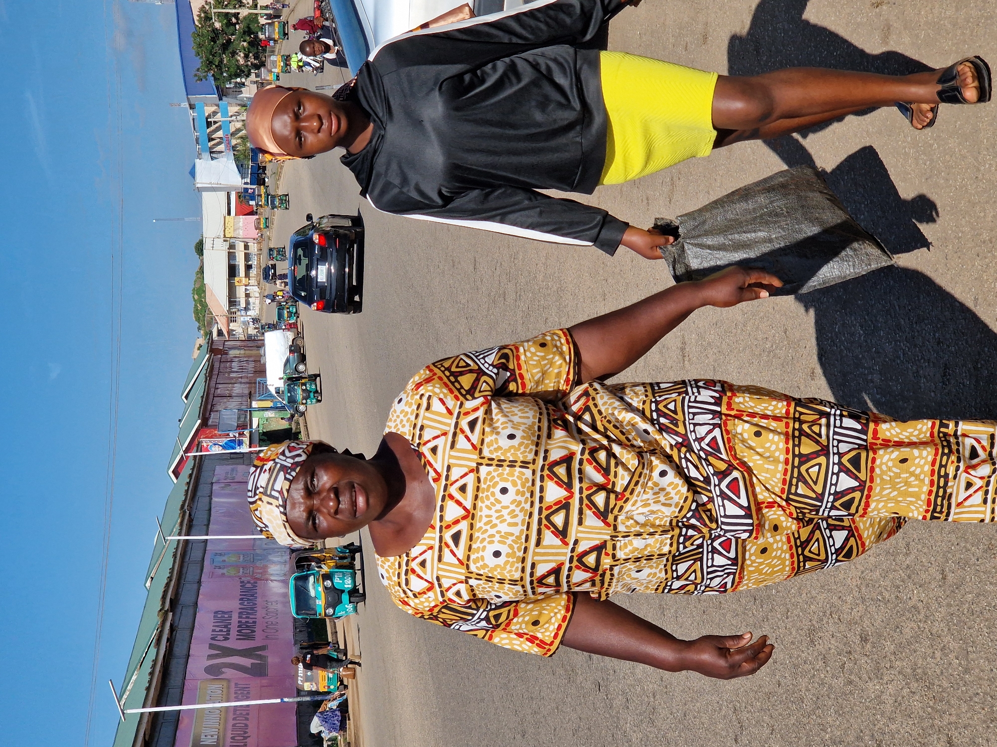In the city of Kabong, Nigeria, on a sunny day, two women walk side-by-side on a paved road. The woman on the left wears a colorful, patterned traditional dress and headscarf. The woman on the right is dressed in a black hooded jacket and a bright yellow skirt, carrying a large grey bag. In the background, several vehicles, including cars and auto-rickshaws, are visible on the road, suggesting a moderately busy urban environment. Buildings line the street, one featuring large pink signage with text that appears to be advertisements. The sky is clear blue.