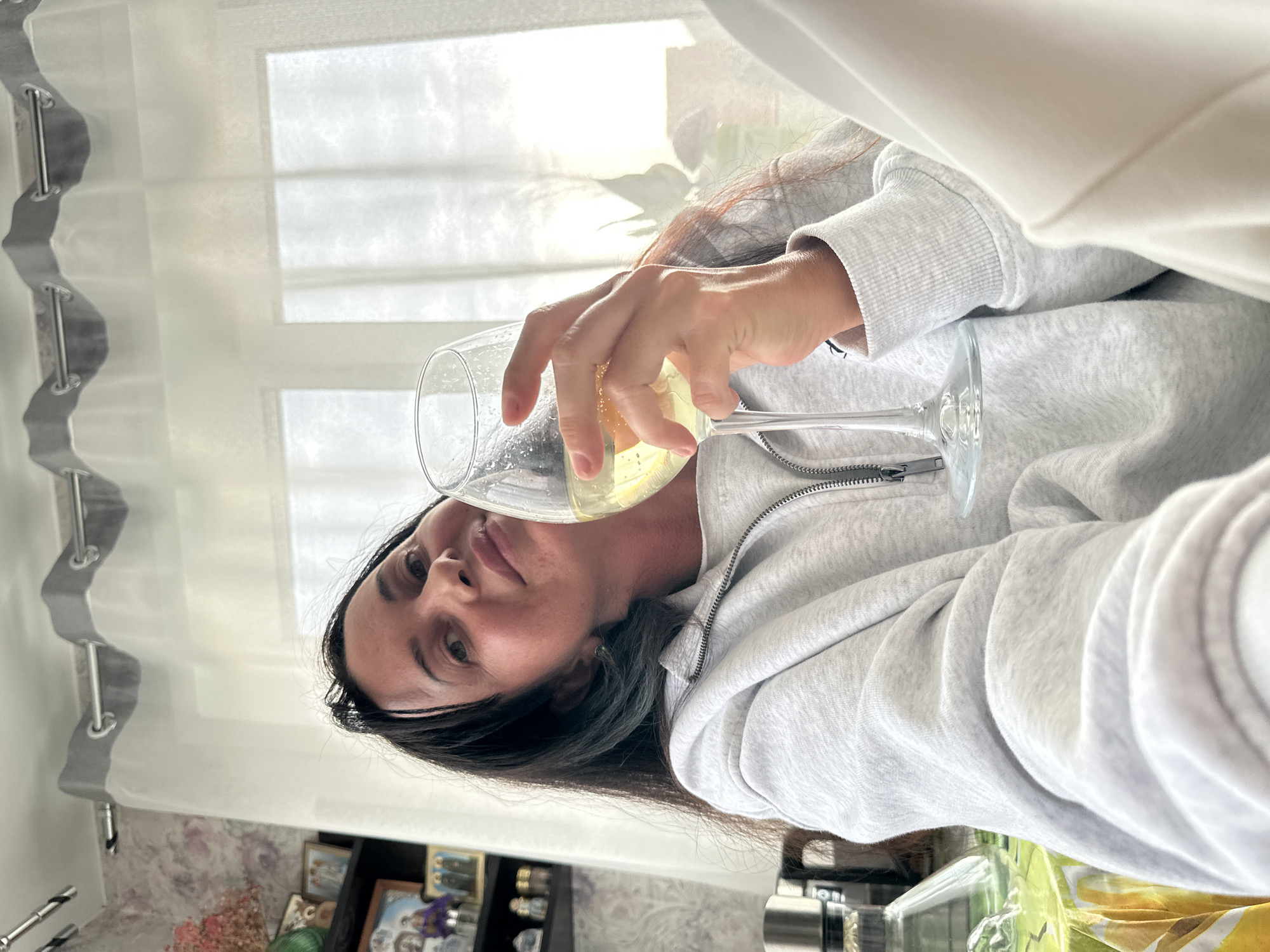A woman with dark hair, wearing a grey hoodie and white pants, is seated indoors, holding a glass of white wine in her right hand. She is looking at the camera with a slight smile. Behind her, sheer white curtains are drawn across a window, with a glimpse of a sunny sky and some foliage outside. To the left of the woman, a shelf with framed photographs and decorative items is visible, and on a table, a glass carafe and other objects are present. The overall setting appears to be a domestic environment, possibly a home in Baklashi, Russia, given the context provided.
