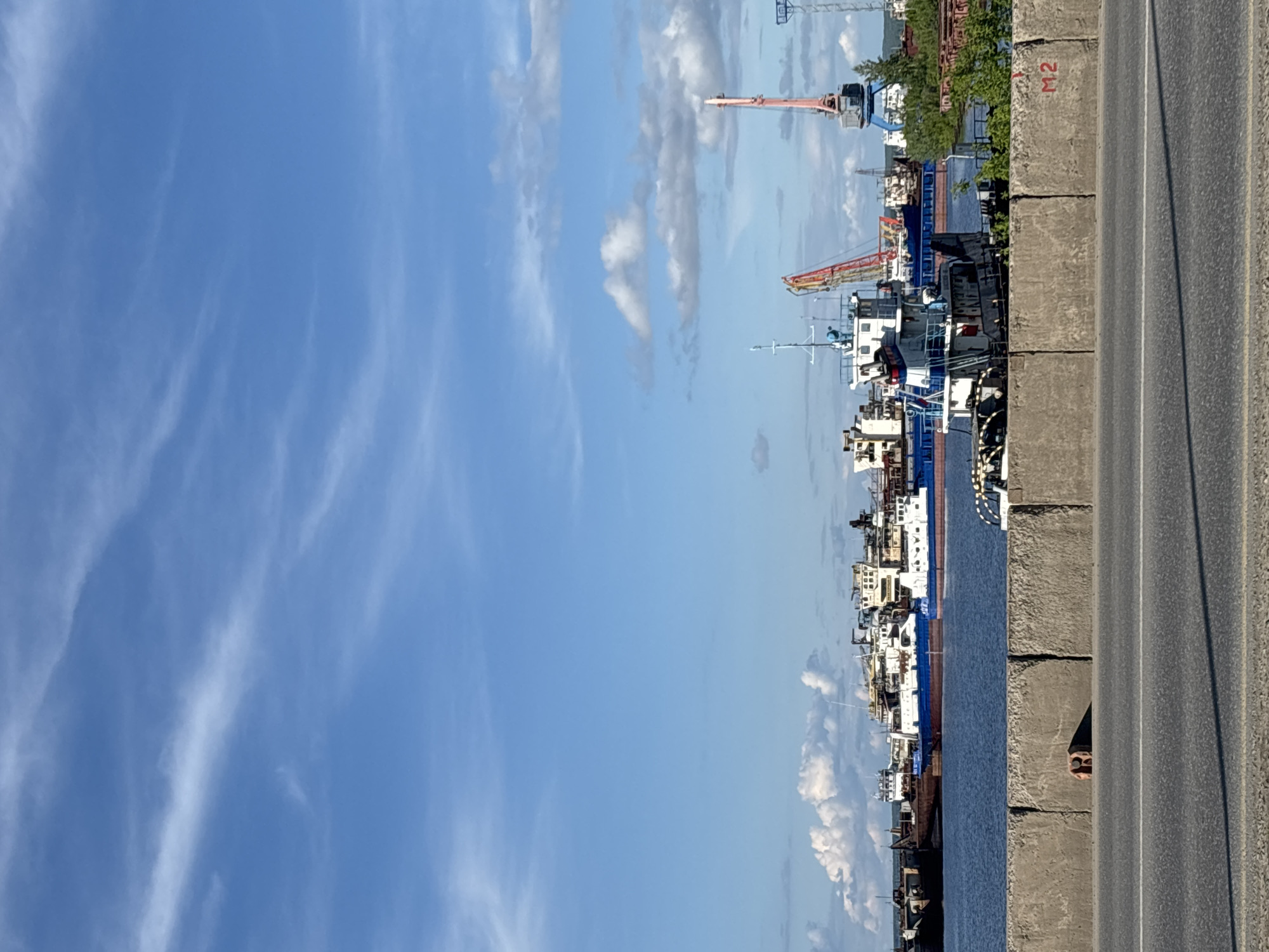 The image depicts a waterfront scene in the city of Chaikovsky, Russia. A concrete barrier separates a road from the water. Several boats, including cargo ships and tugboats, are docked along the shore. Cranes and other industrial equipment are visible in the background. The sky is blue with scattered clouds. There's a person looking out from behind the concrete wall. The environment suggests an industrial port area.