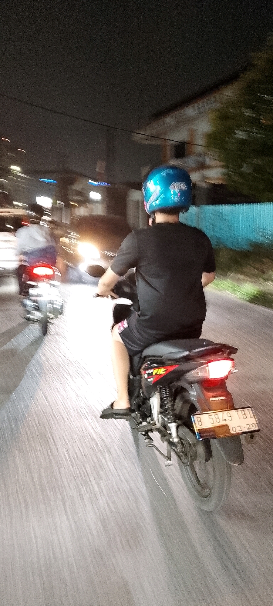 The image, taken at night in Depok, Indonesia, shows two motorcycles on a paved road. The primary subject is a person riding a motorcycle from the rear, wearing a blue helmet with graphics and a black t-shirt and shorts. The motorcycle has a license plate that reads "B 5849 TBI" and a sticker indicating "03-29". The motorcycle also has a red taillight. To the left, another motorcycle is visible, also with a rider, moving in the same direction. The environment appears to be urban, with streetlights and building lights in the background, and trees along the right side of the road. The motion blur suggests the vehicles are in motion.