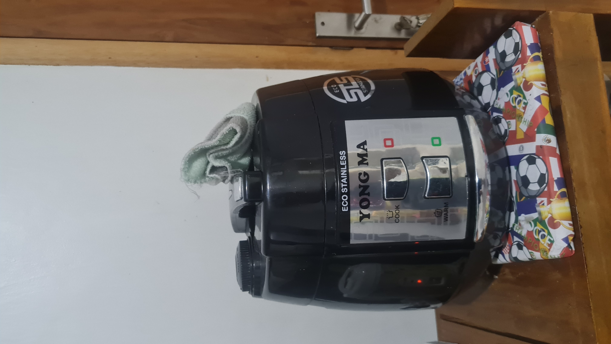 A black Yong Ma brand eco-stainless steel rice cooker is placed on a wooden shelf. The rice cooker has "COOK" and "WARM" indicators with corresponding buttons. A light blue cloth is draped over the top of the rice cooker. The shelf is adorned with a colorful cloth featuring soccer balls and flags. In the background, a white wall and a wooden door frame are visible. The scene is indoors, suggesting a residential setting in Bandar Lampung, Indonesia.