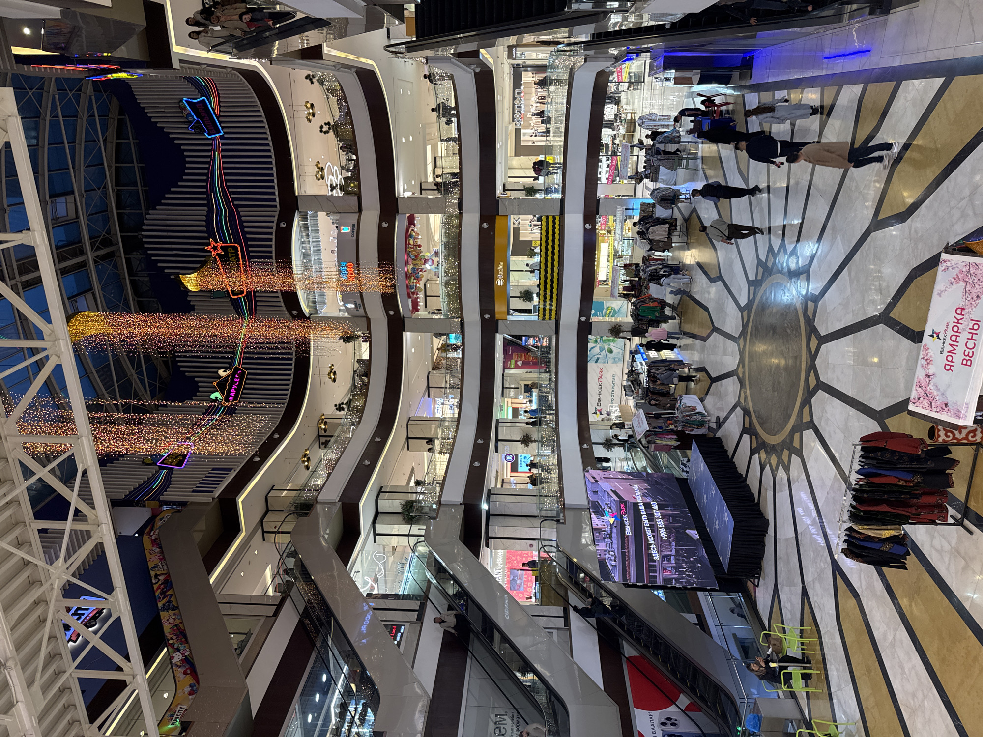 This image displays the vibrant, multi-story interior of a shopping mall atrium, likely BishkekPark in Bishkek City, Kyrgyzstan. The perspective is from an upper level, looking down into the central open space and across multiple floors of retail establishments.
The mall's architecture features several levels of shops with glass facades and dark-railed balconies, all interconnected by numerous escalators visible on either side of the atrium. Overhead, an impressive cascade of warm-toned string lights hangs, creating a sparkling curtain effect. Colorful neon signs, including "КАРУСЕЛЬ" (Carousel), "BISHKEKПАРК," and "Fun CITY," adorn the upper levels, contributing to the lively ambiance.
On the ground floor, a large, dark stage or platform occupies the central area, flanked by a prominent LED screen. The screen displays the mall's name, "BISHKEKPARK," along with an advertisement: "ЗДЕСЬ МОЖЕТ БЫТЬ ВАША РЕКЛАМА" (YOUR ADVERTISING CAN BE HERE) and a contact number (+996 555 307 460). Adjacent to the stage, a sign clearly announces "BISHKEKPARK ЯРМАРКА ВЕСНЫ" (BISHKEKPARK SPRING FAIR), explaining the presence of clothing racks and other merchandise displays on the elaborately patterned marble floor.
Various individuals, mostly adults, are scattered throughout the mall: some are ascending or descending on the escalators, others walk across the ground floor, and a few are seated at tables near the stage. The predominant artificial lighting indicates that the scene is set during the evening or night. Visible shop names include "Benline," "COFFEE," and "MARK." Other legible text includes "БААЛАР!" (prices) and "ПРО ОТКРЫТИЕ!" (about opening). The overall atmosphere is one of a bustling, modern shopping center hosting a special event or fair.