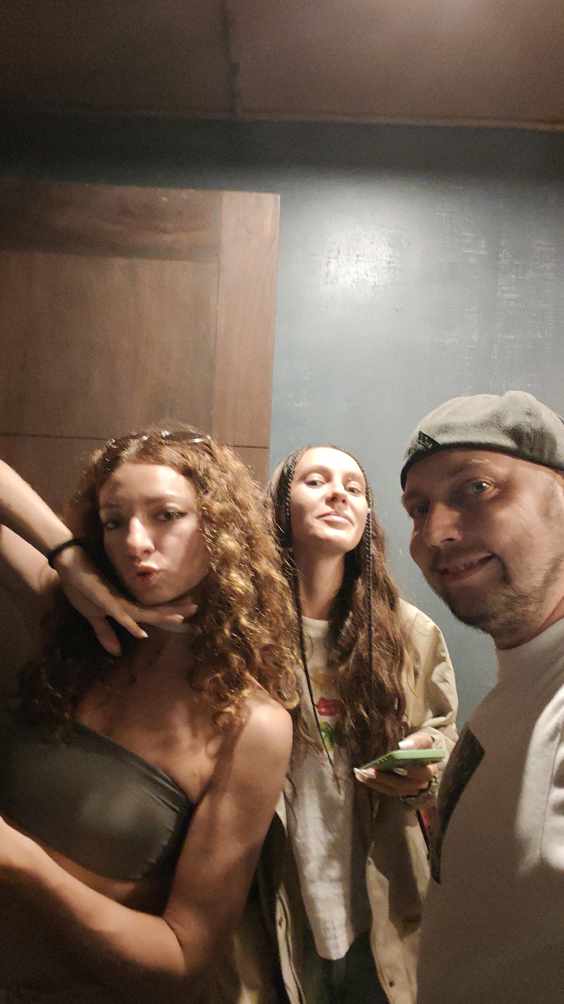 The image depicts an indoor scene in Mu Cang Chai, Vietnam, featuring three individuals. On the left, a female with long, curly reddish-brown hair, wearing a dark olive-green strapless top, poses with a pout while looking towards the camera. Her chin is gently cupped by the hand of the adjacent female. This second female, positioned centrally, has long, dark hair styled in thin braids, wears a light long-sleeved jacket over a graphic t-shirt, and holds a green smartphone in her right hand while looking slightly upwards with a faint smile. To the far right, a male with a short beard, wearing a grey baseball cap backward and a light t-shirt, smiles broadly at the camera. The background consists of a distinct interior corner, with a muted brownish-orange paneling on the left and a textured bluish-gray wall on the right. The lighting is dim, and the image exhibits a grainy texture.