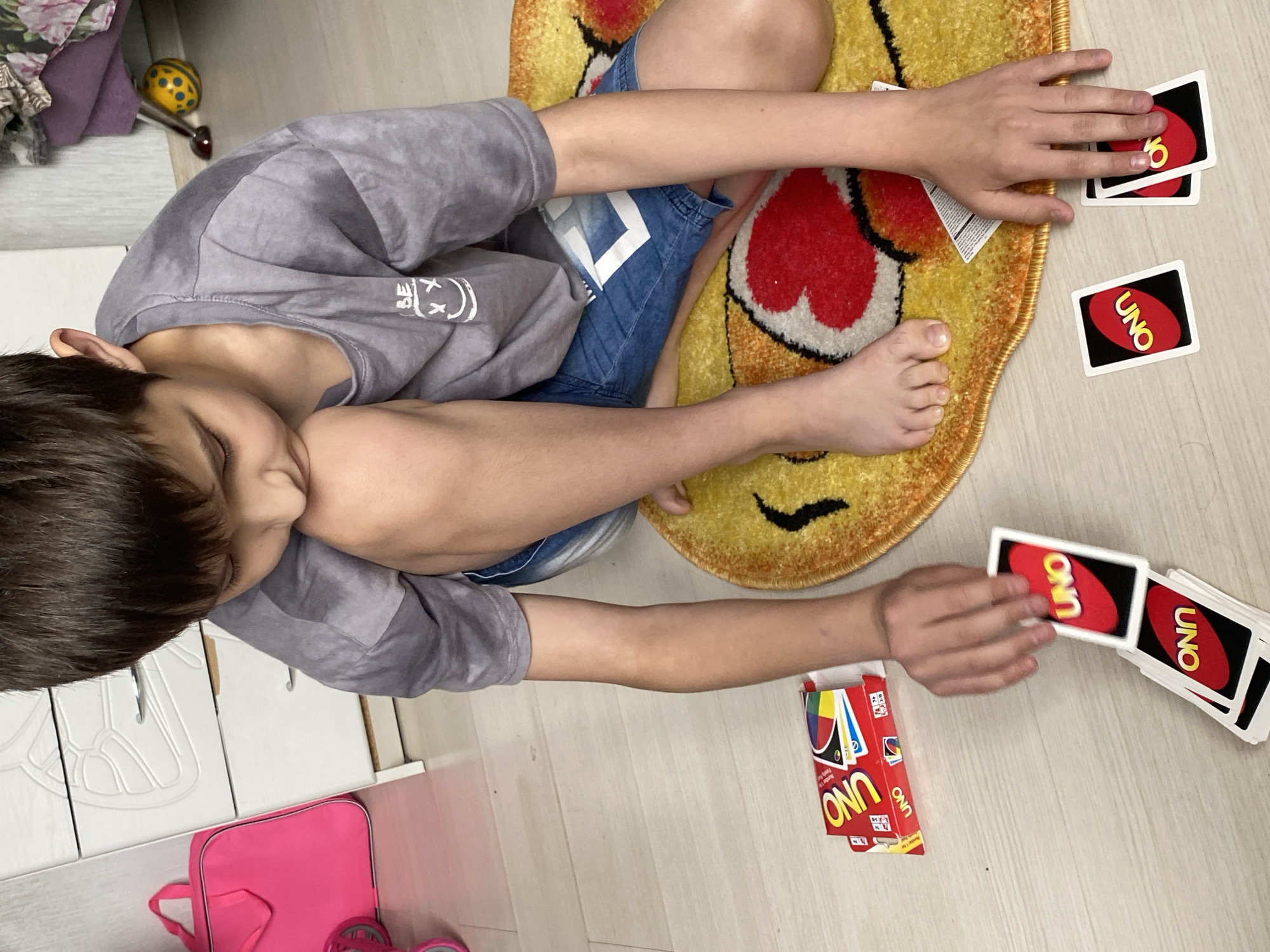 An overhead shot captures a young person, likely a child, playing a card game on a light wooden floor in Chaikovsky, Russia. The person is lying on their left side, with their head resting on their left arm, and their body partially on a round, yellow, fuzzy rug with red and black accents, possibly depicting a character like Pikachu. They are wearing a grey short-sleeved t-shirt with a white graphic on the back and blue denim shorts.
Their right arm is extended towards a stack of "UNO" cards. One hand is holding three cards, two of which clearly show the "UNO" logo in red and yellow on a black background. Another two "UNO" cards are visible separately on the floor, one showing a red "6" and the other with the "UNO" logo. An empty "UNO" card box, predominantly red with colorful game card illustrations, is also on the floor near their right hand.
In the upper left of the frame, there's a glimpse of a cluttered area with what appears to be folded fabric, including purple material, and a yellow polka-dotted ball. In the lower left, a bright pink bag is visible, suggesting other belongings are nearby. The lighting suggests it's daytime indoors. The child's facial expression is not clearly visible as their head is turned away.