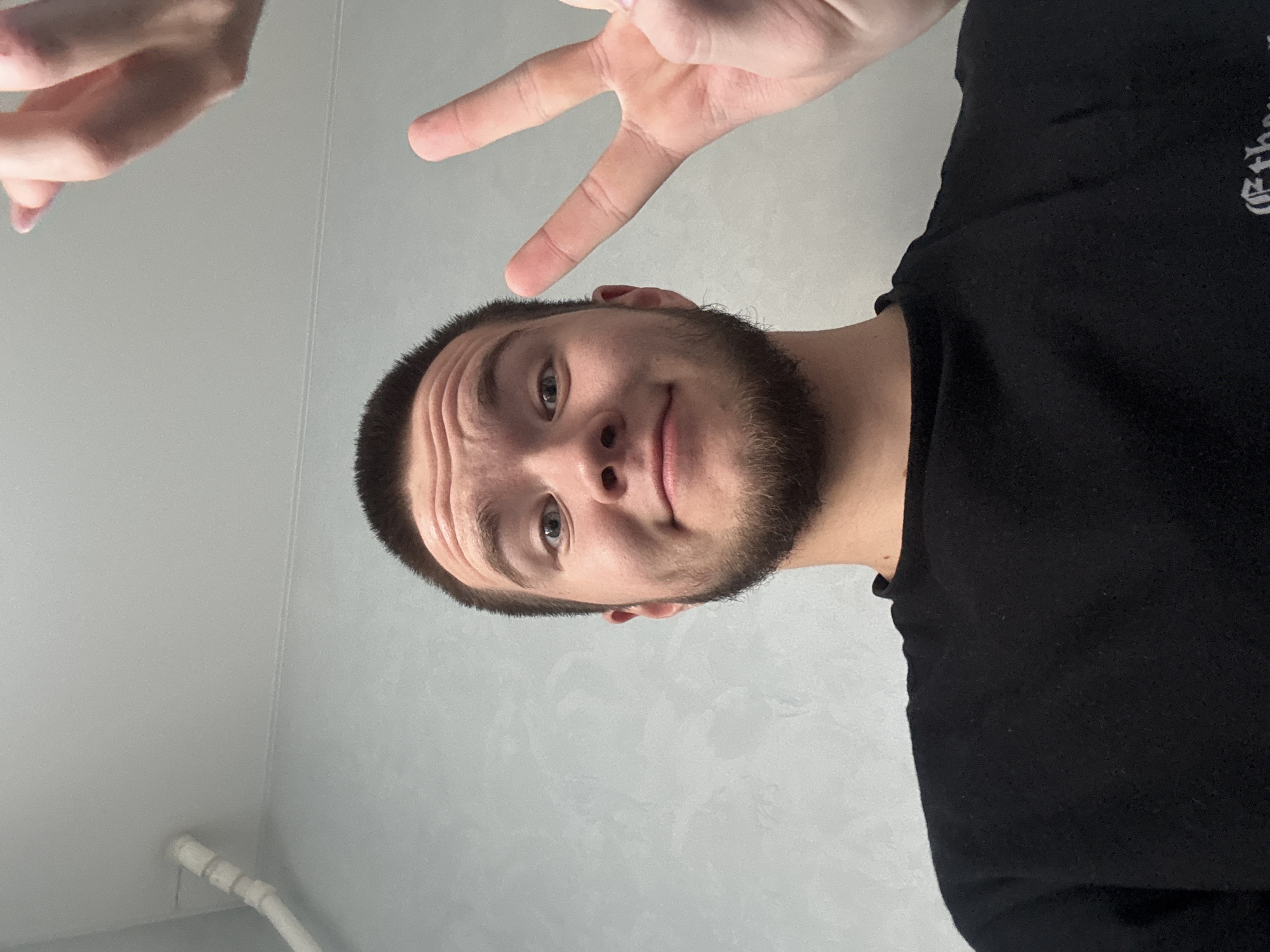 A man with short dark hair and a beard is looking at the camera with a slight smile. He is wearing a black t-shirt and making a peace sign with his left hand above his head. The background is a light gray wall with a white pipe running along the bottom. The location appears to be indoors, likely a residential building. The location in the city of Sarapul, Russia, cannot be confirmed from the image alone.