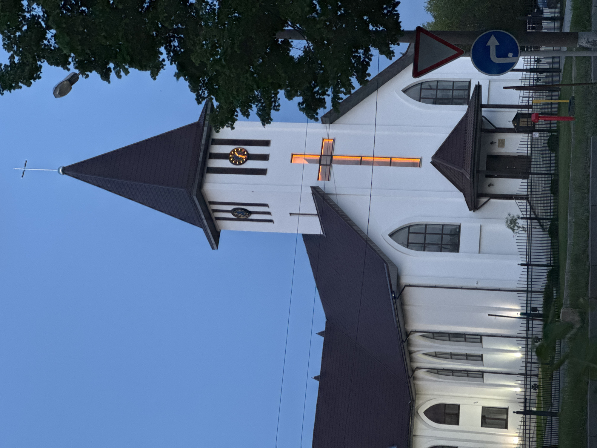 The image shows a white church with a brown roof against a blue sky. The church has a tall tower with a clock and an illuminated cross. A tree is visible in the upper portion of the image. In the foreground, there is a traffic sign. A fence surrounds the church grounds. The location is in an urban setting, in the city of Unknown, Unknown.