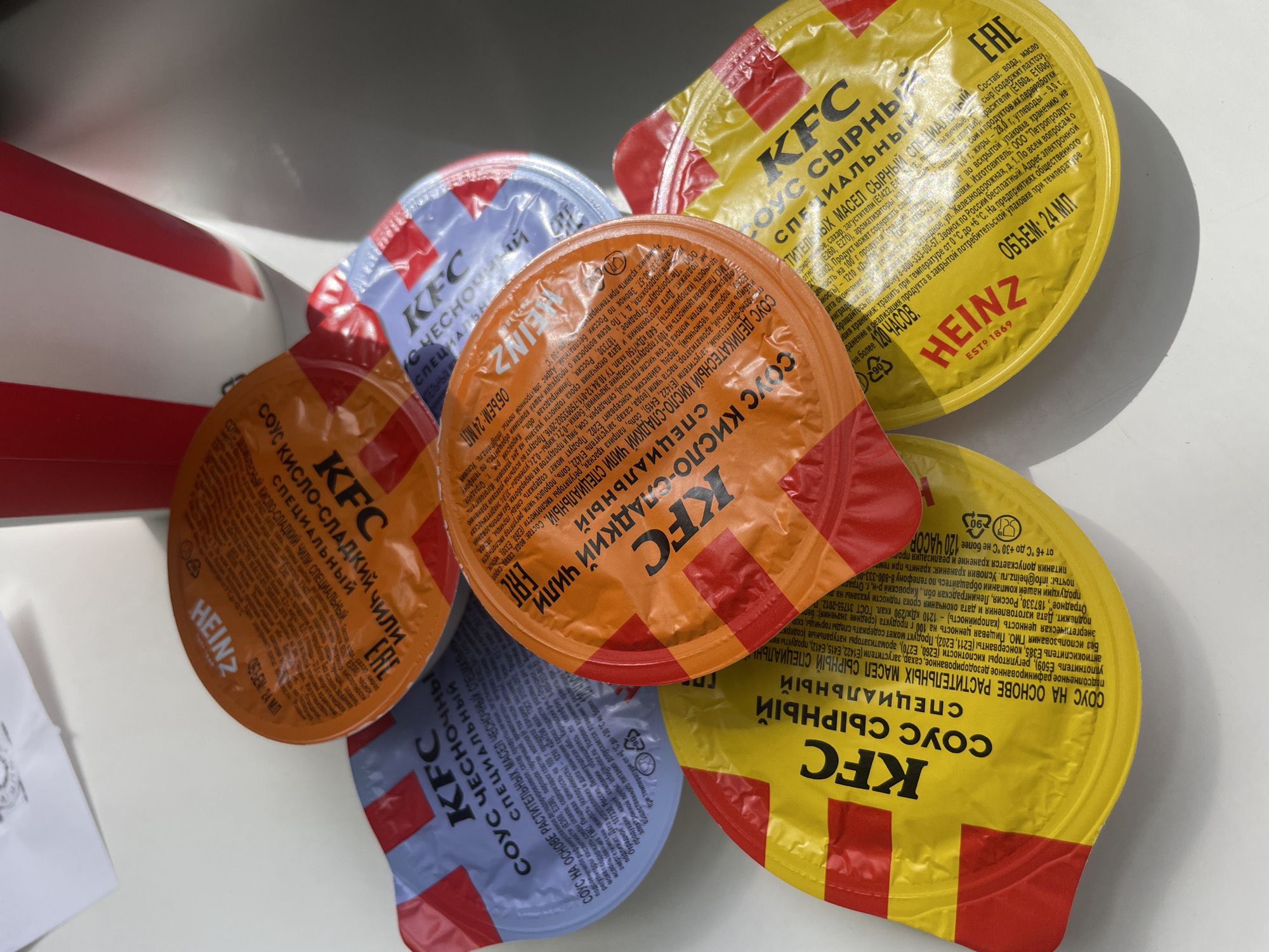The image shows a collection of KFC sauce packets. There are several varieties visible, including 'Cheese', 'Sweet and Sour Chili', and 'Garlic'. The packets appear to be made of foil with printed labels in Russian. The labels include the KFC logo, flavor description, ingredient information, and volume (24 ml). The packets are resting on a light surface. A small portion of a KFC container is visible in the background. The location in Yerevan, Armenia, is unknown.