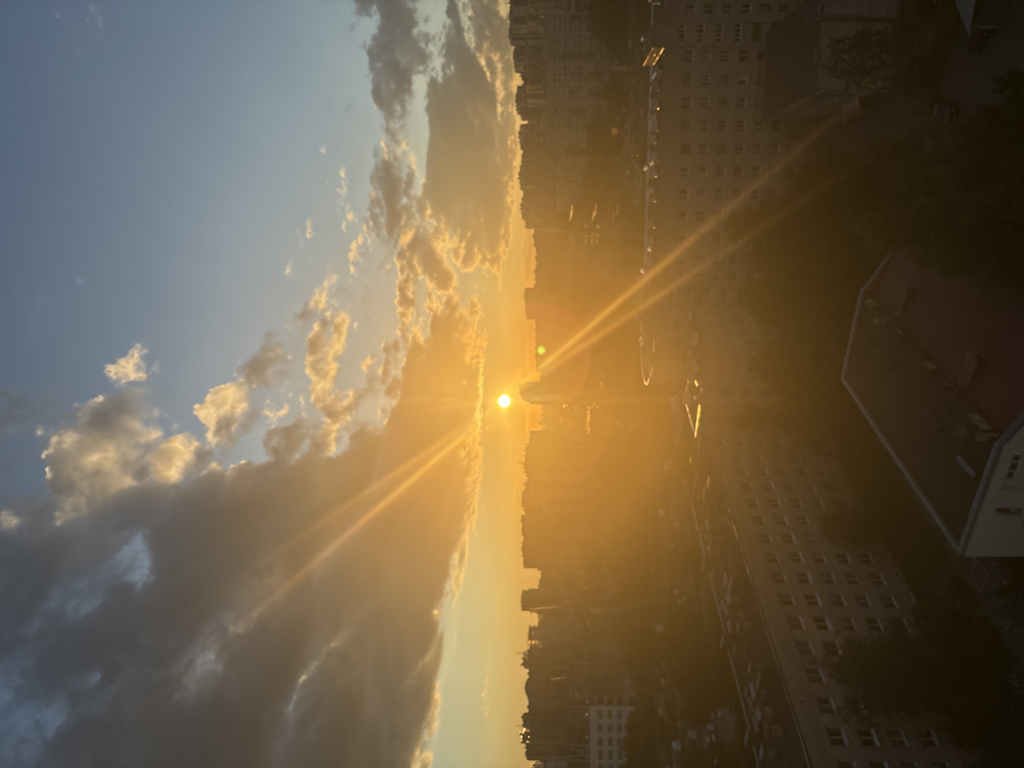 This is a high-angle, full shot of a city skyline at sunset. The sun is low on the horizon, casting a warm golden glow over the scene. Large, dramatic clouds are present in the sky, some tinged with orange and pink from the setting sun. The city itself is densely packed with buildings of varying heights, appearing as silhouettes against the bright sky. In the foreground, several apartment buildings are visible, with rows of windows. Trees and other greenery can be seen interspersed between the buildings. The overall impression is one of an urban environment bathed in the soft, atmospheric light of a sunset.
