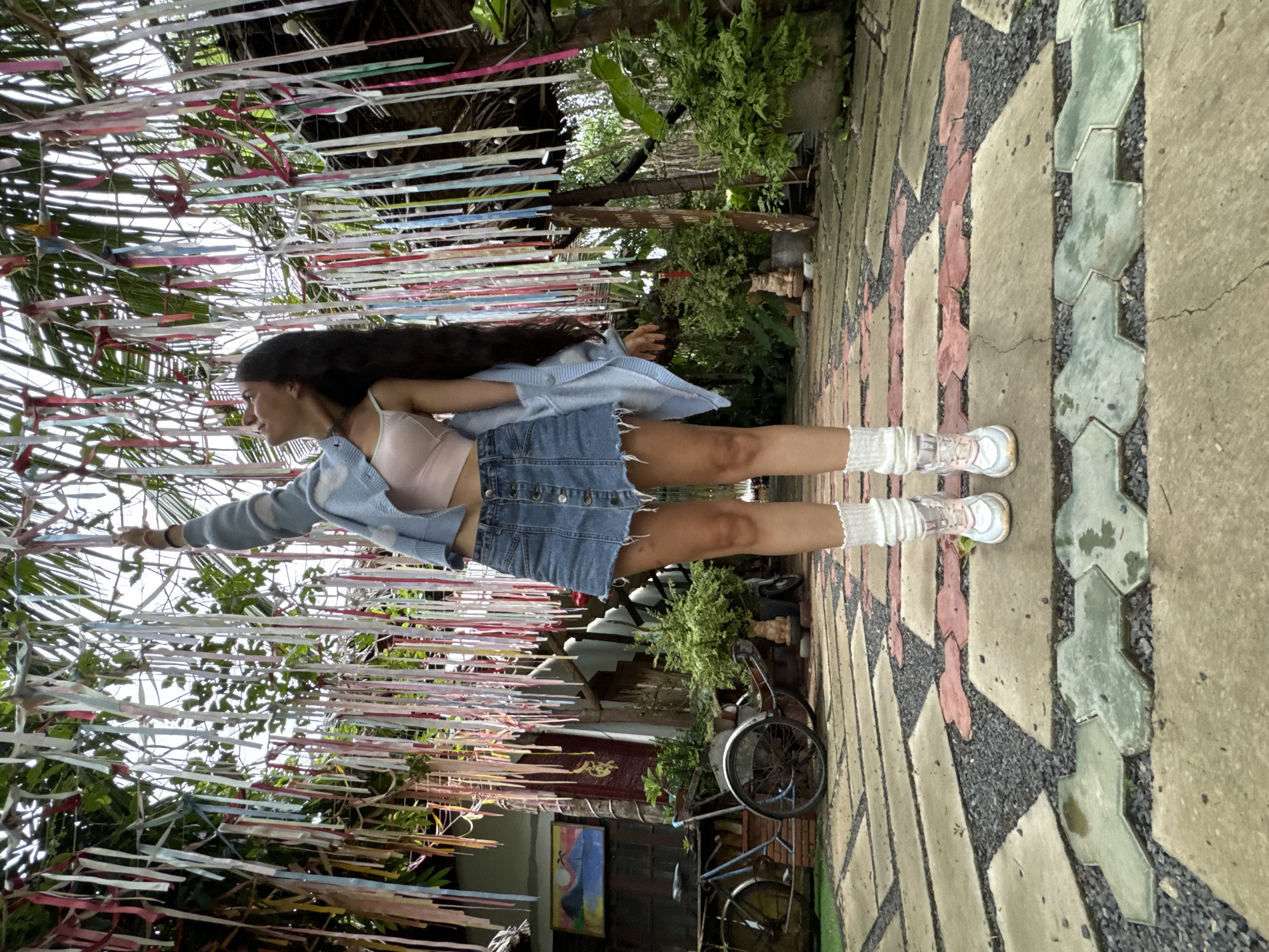 A full shot captures a woman standing in what appears to be an outdoor setting. She is wearing a light pink tank top, a denim skirt, a light blue button-down shirt, white socks, and shoes. Her hair is long and dark. She stands on a stone-paved area with geometric designs. To her left are numerous hanging ribbons of various colors, creating a tent-like effect. Behind her, there's a glimpse of foliage and plants, suggesting a garden or natural environment. In the background, one can spot a bicycle. The image is taken at a slightly unusual angle. Given the city mentioned, this could be a tourist location or an outdoor space in Mũi Né, Vietnam.