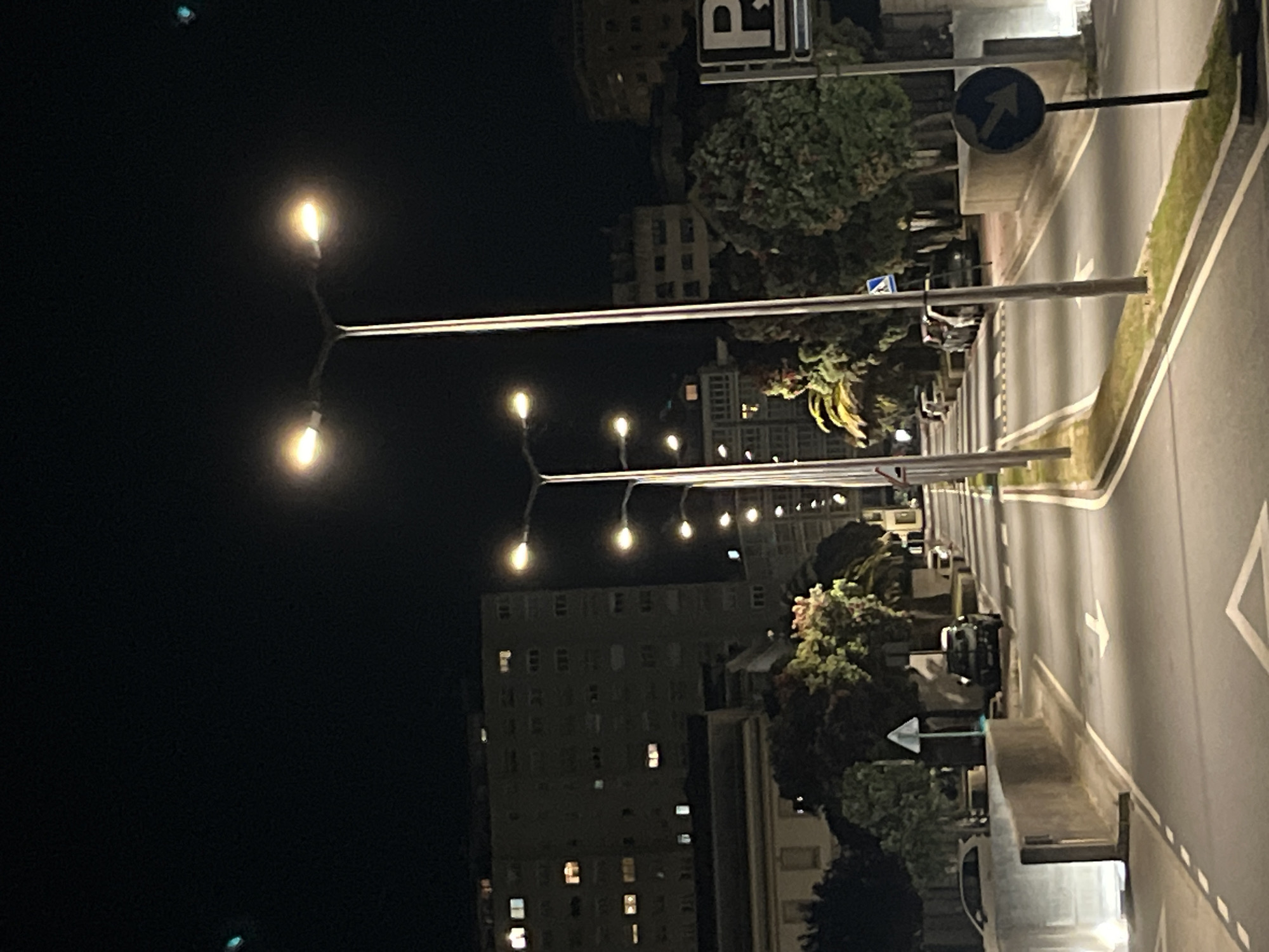 The image depicts a night scene in A Coruña, Spain. A street illuminated by a series of old-style streetlights runs alongside a row of buildings. A blue arrow road sign is visible, indicating the direction of traffic. Several parked cars line the side of the street. The buildings appear to be residential, with lit windows suggesting occupancy. The sky is dark.