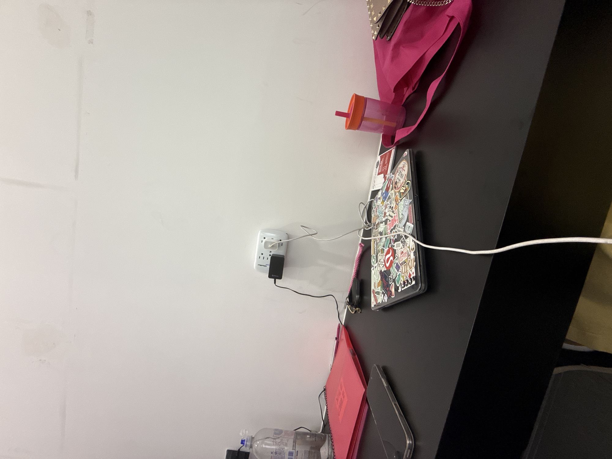 The image depicts a scene in San Bernardino Tlaxcalancingo, Mexico. A white wall is visible on the left side of the frame. An electrical outlet with a black adapter plugged in is attached to the wall. A black desk is seen on the right, on which several items are placed. These items include a pink bag, a pink cup with an orange lid, a phone case covered in stickers, a pink notebook with a spiral binding, a smartphone, and a clear water bottle. A white cable runs from the phone case to the electrical outlet.