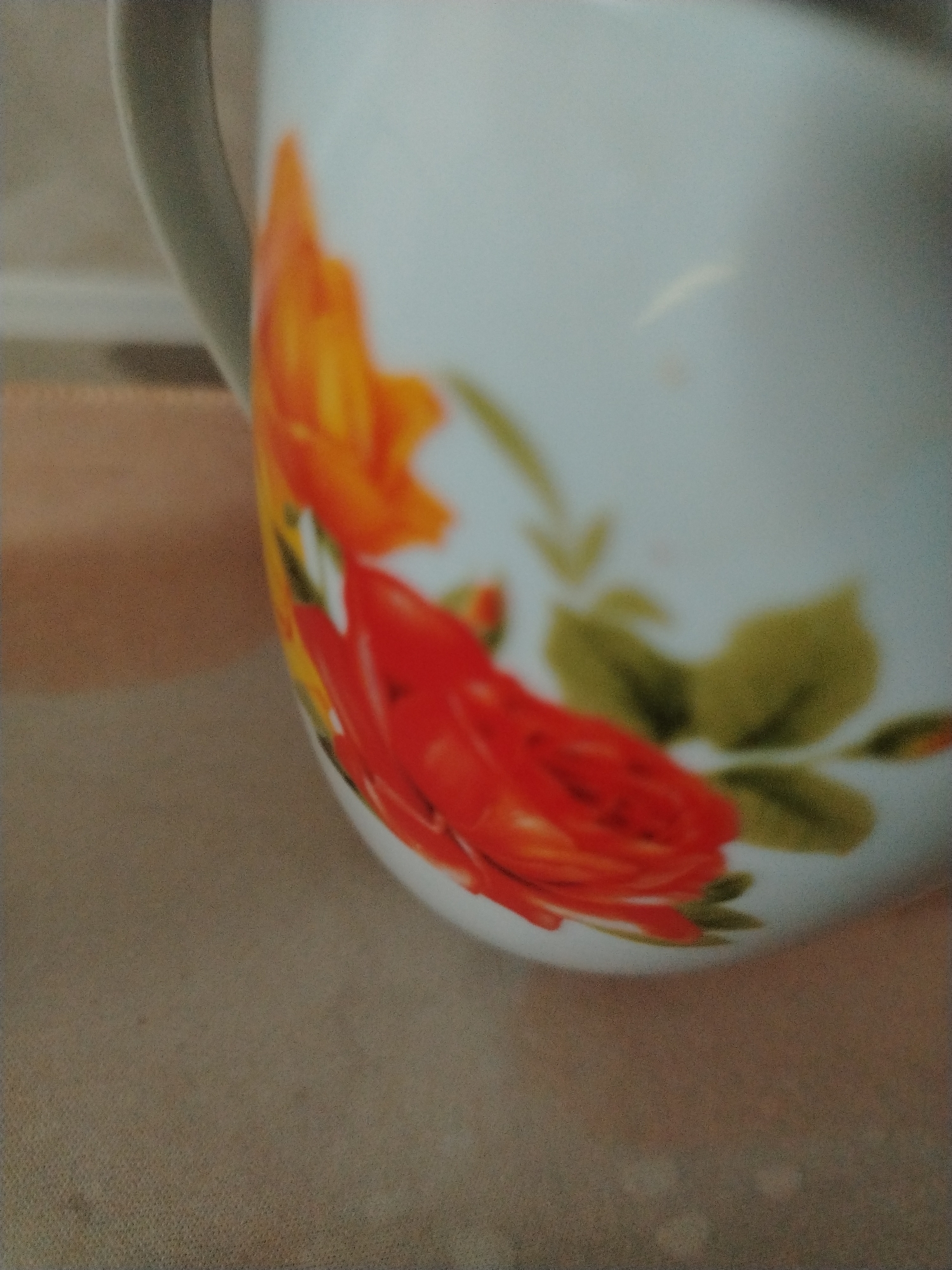 The image shows a close-up of a white ceramic pitcher with a floral design. The pitcher has a handle on the left side. The floral design features red and orange roses with green leaves. The pitcher is placed on a textured surface that appears to be a table or countertop. The background is blurred and out of focus, suggesting a shallow depth of field. The lighting is soft and diffused, highlighting the colors and textures of the pitcher and its design.