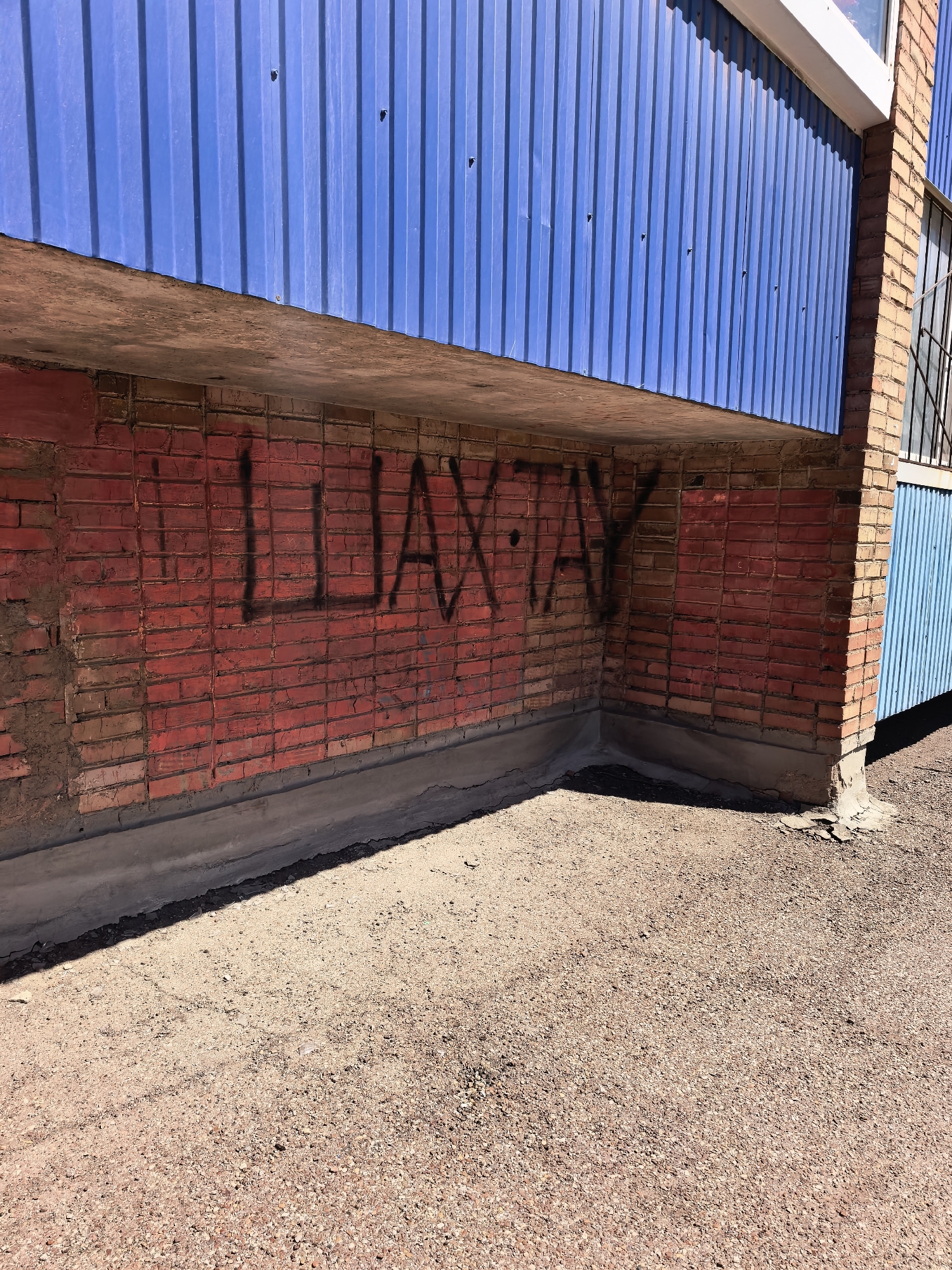 A low-angle, eye-level shot depicts a brick building with black graffiti under a blue corrugated metal overhang. The graffiti appears to spell "ШАХТАУ" in Russian. The building is constructed of reddish-brown bricks, and the base of the building is concrete. The overhang is a light blue corrugated metal. Above the overhang is another portion of the building with a window and metal bars. The ground is covered in reddish-brown gravel. The location is Sterlitamak, Russia.