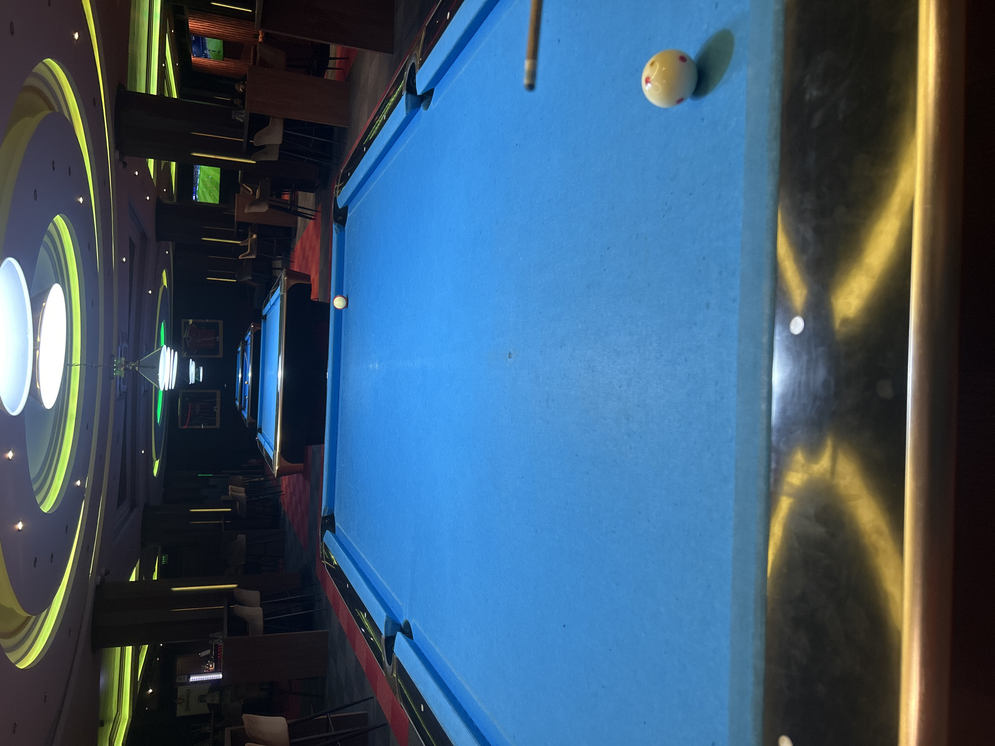 The image shows the interior of a bar or entertainment establishment featuring pool tables. The location is potentially Sofia, Bulgaria, though no specific details in the image confirm this. The main subject is a blue-felt pool table with visible cue stick and cue ball in the foreground. Another pool table with a white ball is visible in the background. The environment is dimly lit with decorative ceiling lights and surrounding bar furniture including tables and chairs. There is no evidence of any people.