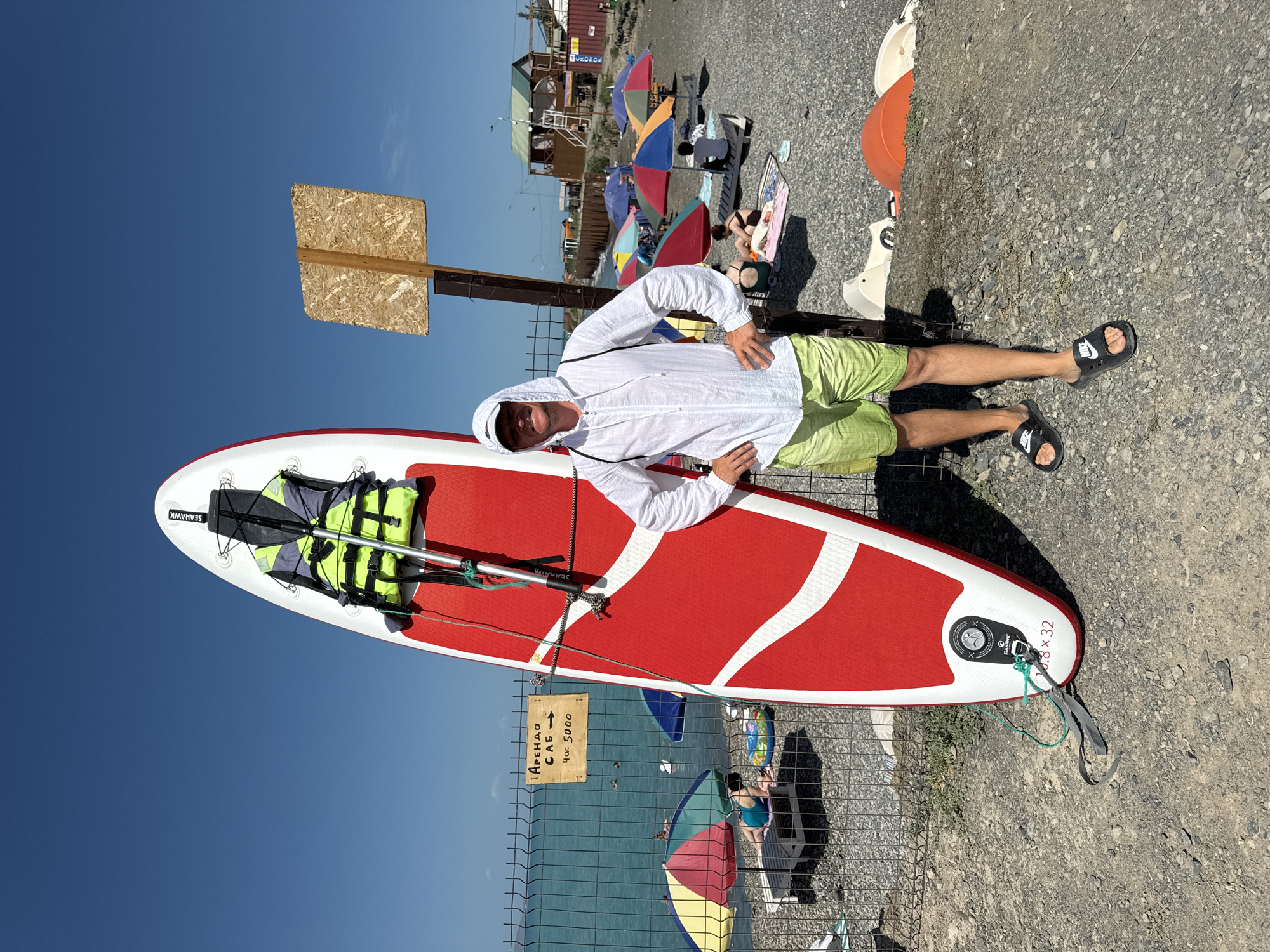 A man in a white hooded jacket and green shorts stands on a rocky beach next to a red and white stand-up paddleboard. The paddleboard has a yellow and green life vest attached to it. Behind the man, beach umbrellas and lounge chairs are set up. The sea is visible in the background, with a few people swimming. A wooden sign with Cyrillic text is posted on a fence to the left of the man. This scene is located on the shore of Lake Zharbulak in Zharbulak, Kazakhstan.